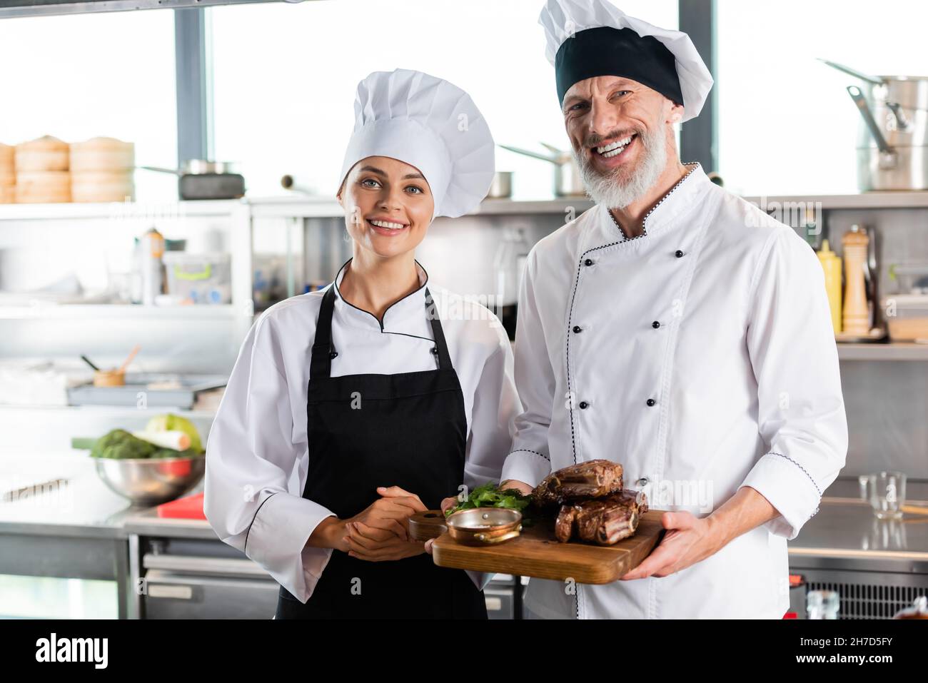 Supportive Chefs Positive chefs smiling at camera while holding roasted meat in kitchen ...