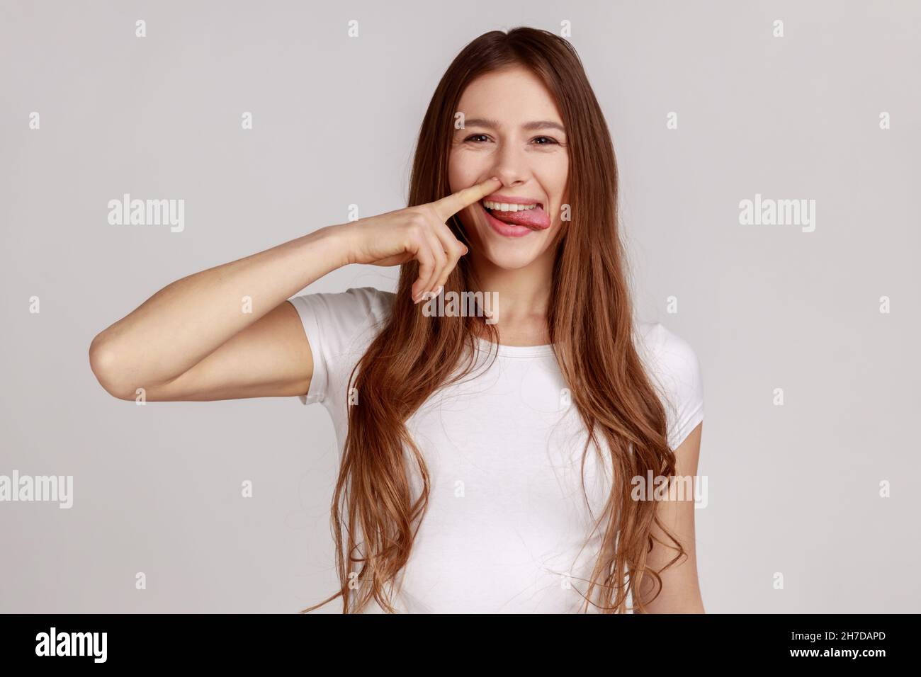 Funny cheerful woman picking nose with stupid silly face, pulling out boogers, bad manners concept, misconduct, wearing white T-shirt. Indoor studio shot isolated on gray background. Stock Photo