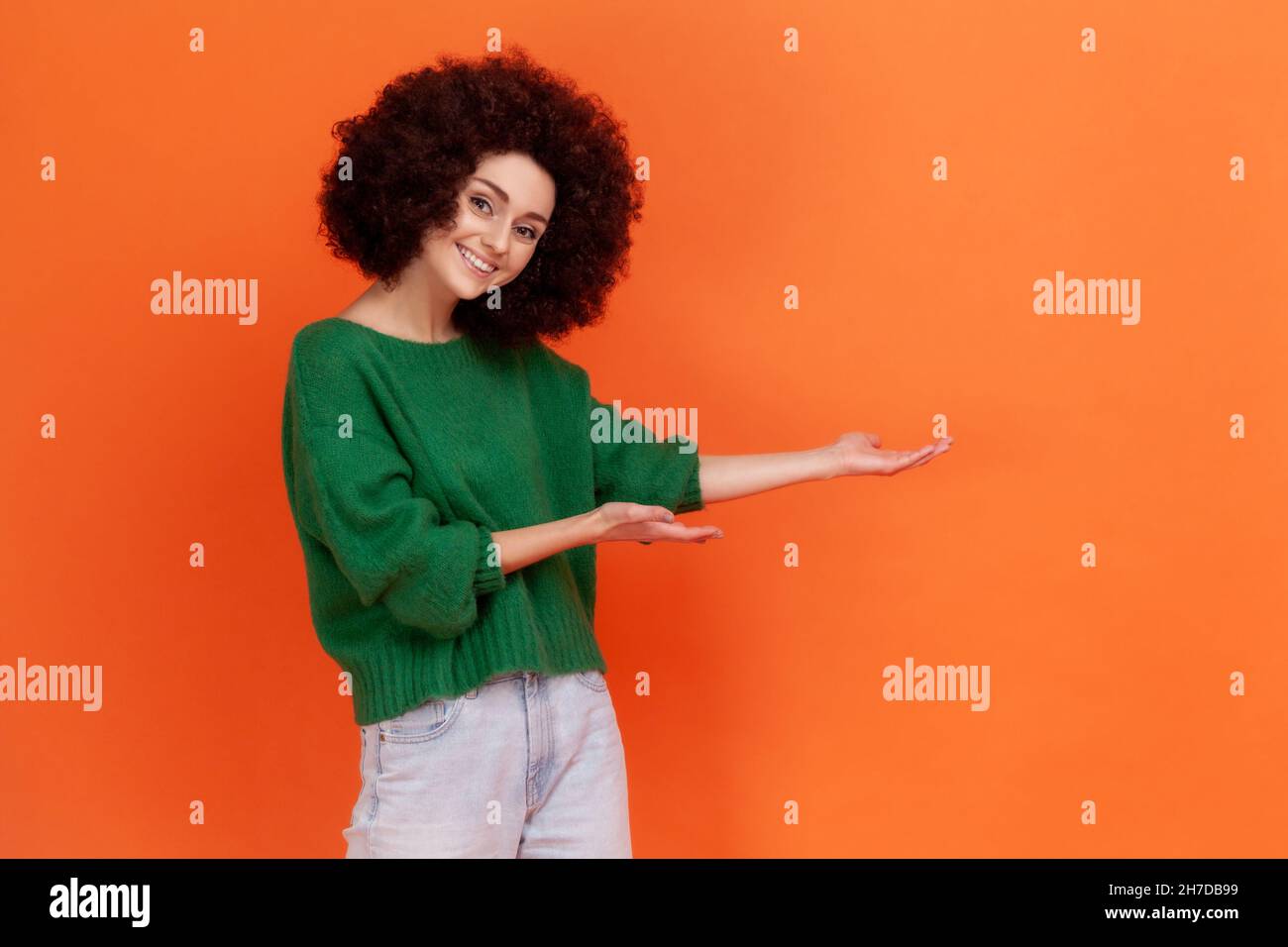Happy friendly woman with Afro hairstyle wearing green casual style sweater presenting advertising area with toothy smile, copy space. Indoor studio shot isolated on orange background. Stock Photo