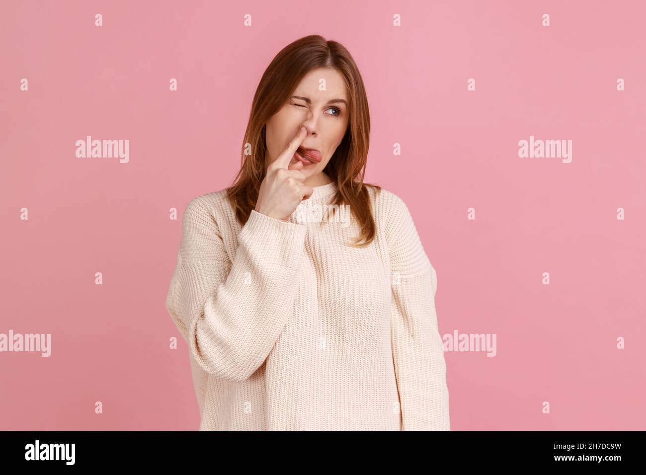 Portrait of blond woman sticking out tongue while picking nose, looking at camera with foolish silly expression, wearing white sweater. Indoor studio shot isolated on pink background. Stock Photo