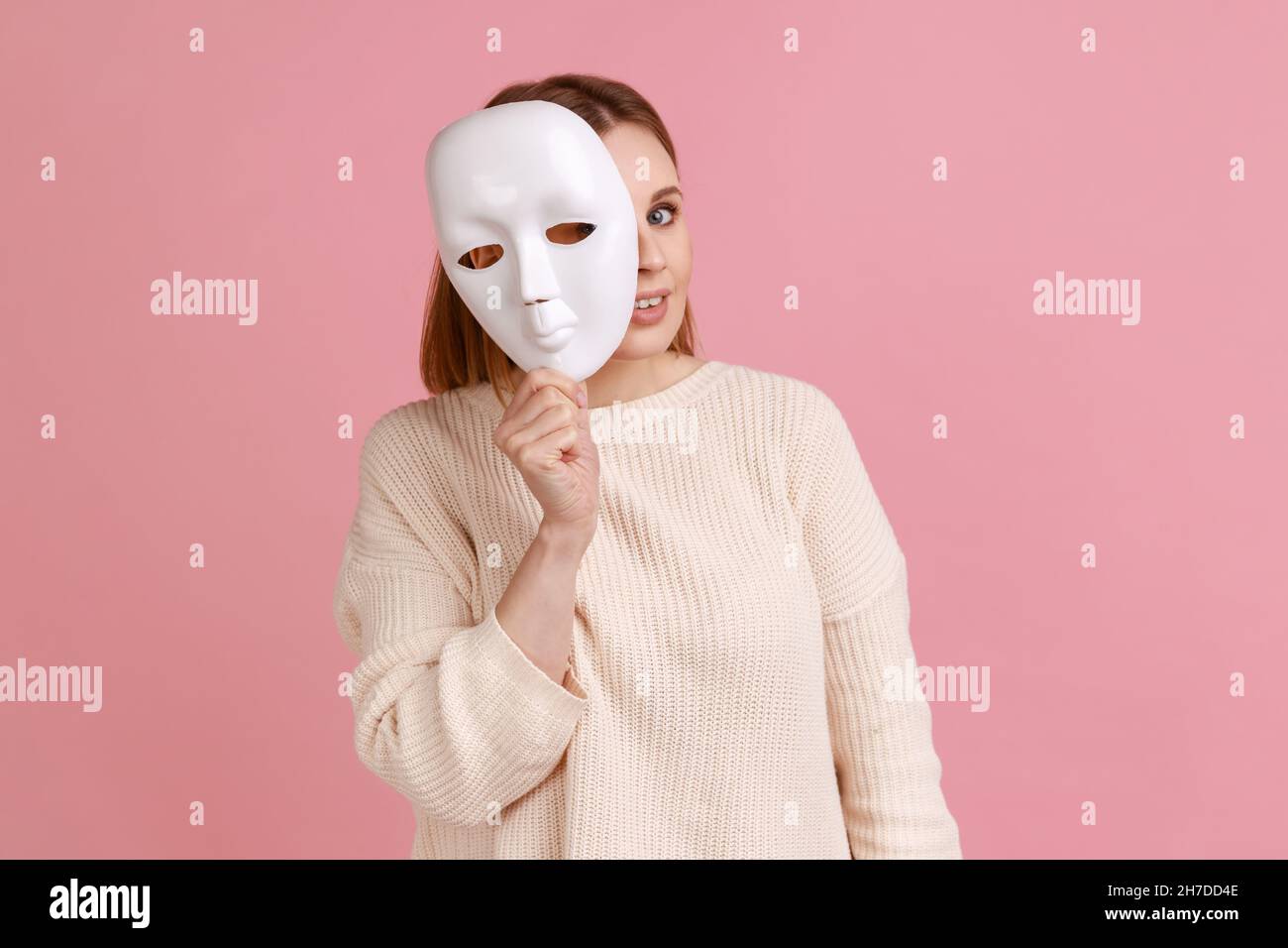 Portrait of smiling blond woman holding white mask, peeking, looking at camera, pretending to be another person, wearing white sweater. Indoor studio shot isolated on pink background. Stock Photo