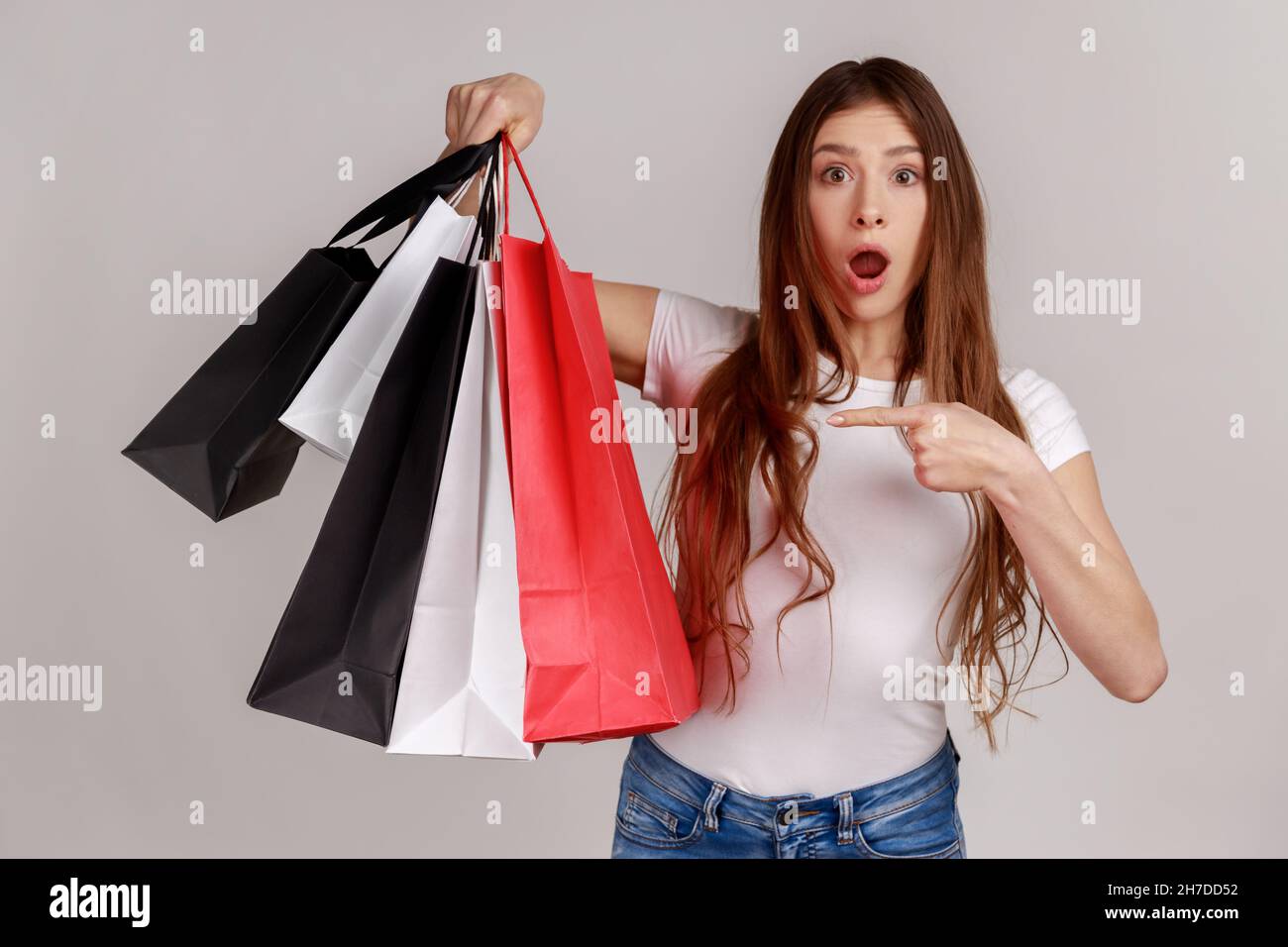 Shocked amazed dark haired woman pointing finger at paper bags in her hand, surprised with shopping, low prices good quality, wearing white T-shirt. Indoor studio shot isolated on gray background. Stock Photo