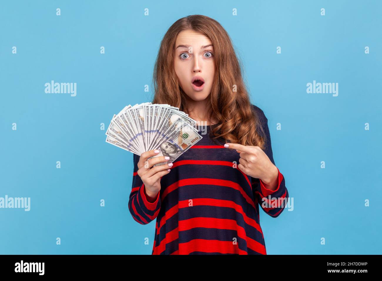 Shocked woman wearing striped casual style sweater, pointing at dollars banknotes in her hand, having astonished expression, big profit. Indoor studio shot isolated on blue background. Stock Photo