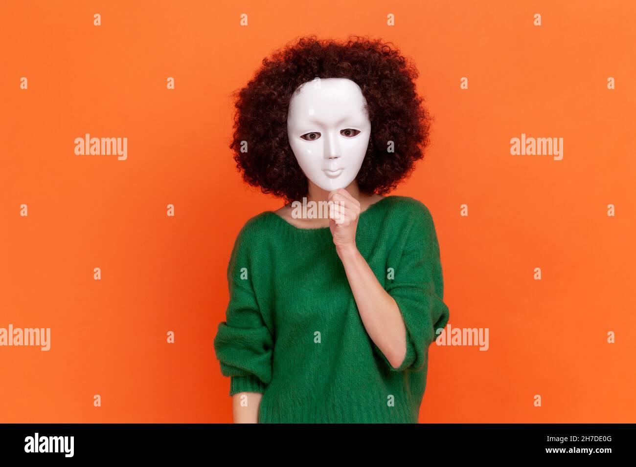 Unknown woman with Afro hairstyle wearing green casual style sweater hiding her face behind white mask, hiding her personality, anonymous. Indoor studio shot isolated on orange background. Stock Photo