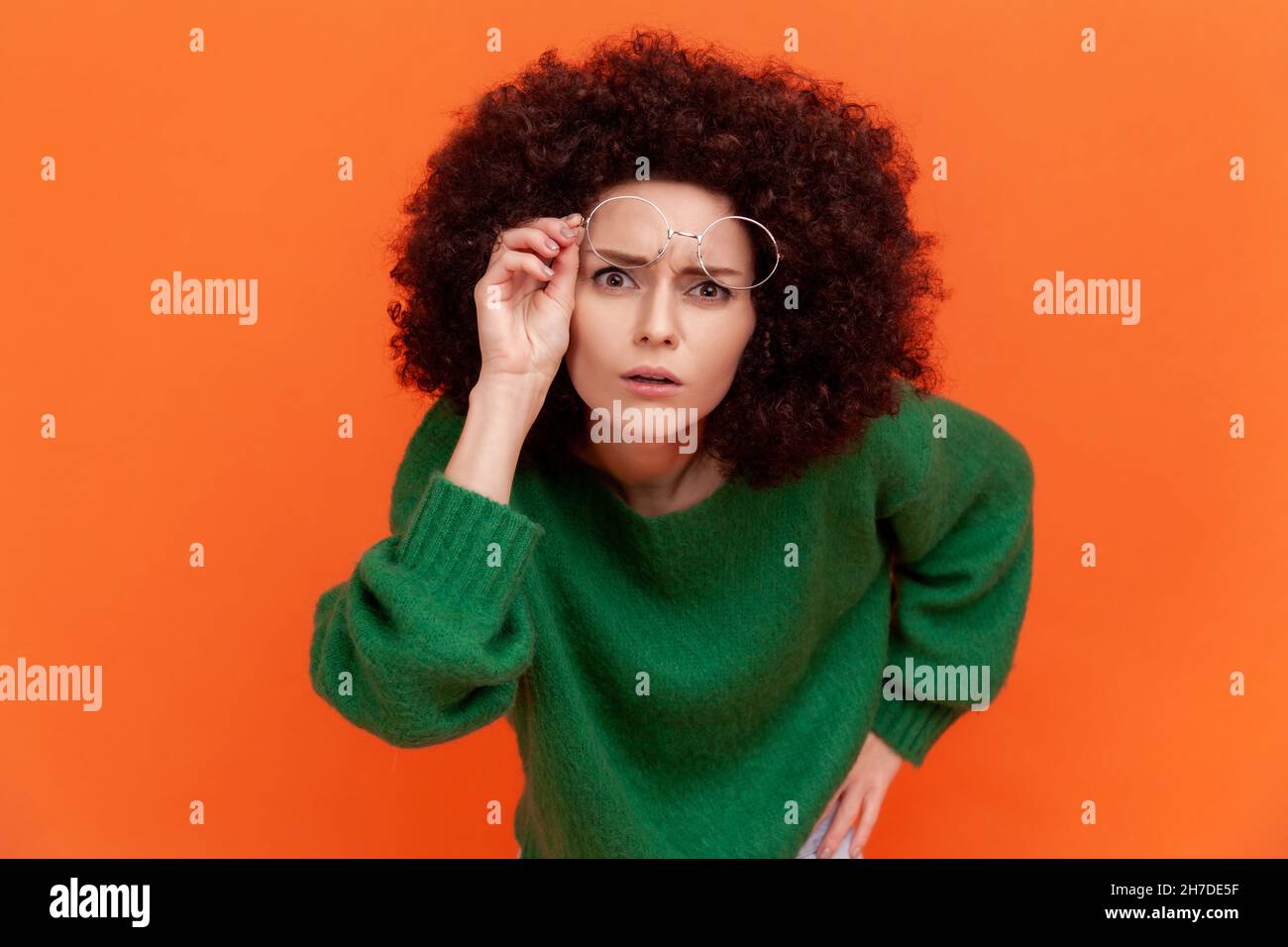 Curious woman with Afro hairstyle wearing green casual style sweater raising her optical glasses, wants to see something better, staring attentively. Indoor studio shot isolated on orange background. Stock Photo