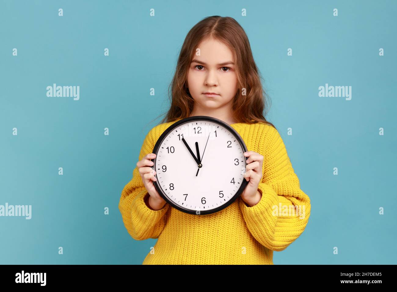 Portrait of little girl holding big wall clock in hands, looking at camera with serious expression, wearing yellow casual style sweater. Indoor studio shot isolated on blue background. Stock Photo