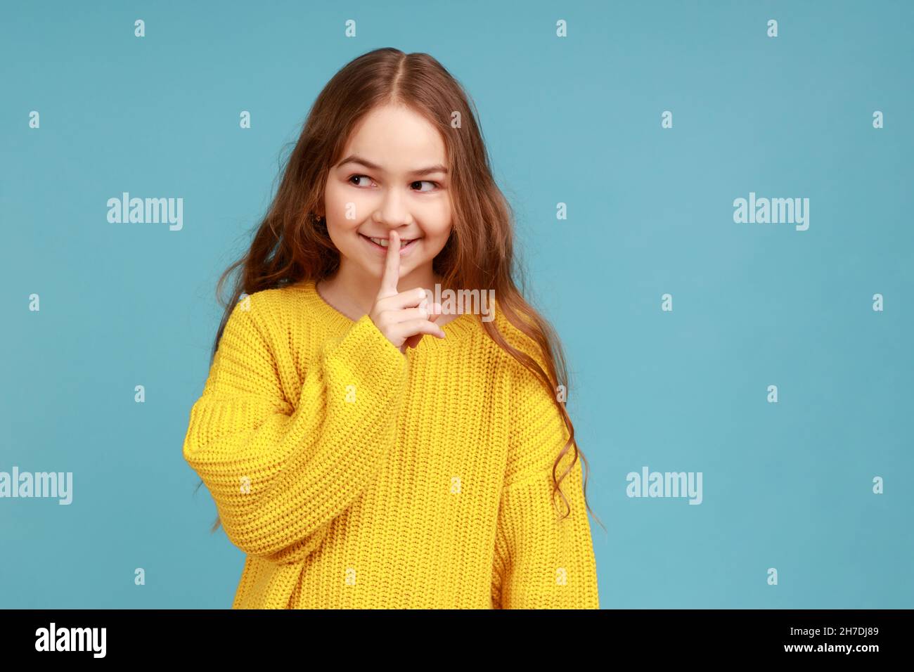 Portrait of adorable little girl making silence gesture with finger on lips, looking away with smile, wearing yellow casual style sweater. Indoor studio shot isolated on blue background. Stock Photo