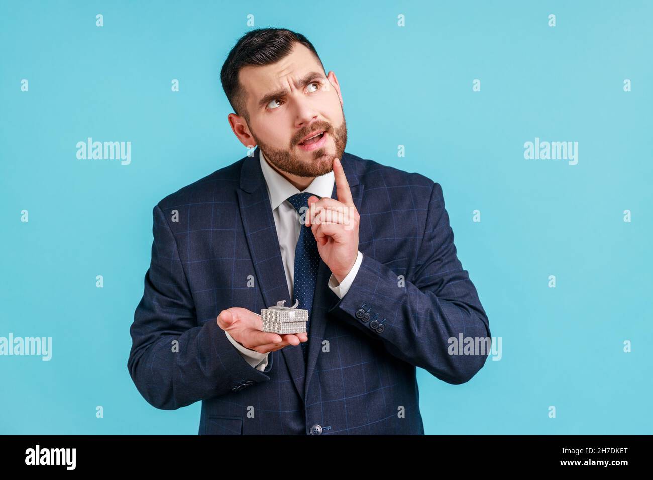 Portrait of pensive man with beard wearing official style suit holding in hands small box with ring, thinking the way to make proposal to his girlfriend.Indoor studio shot isolated on blue background. Stock Photo