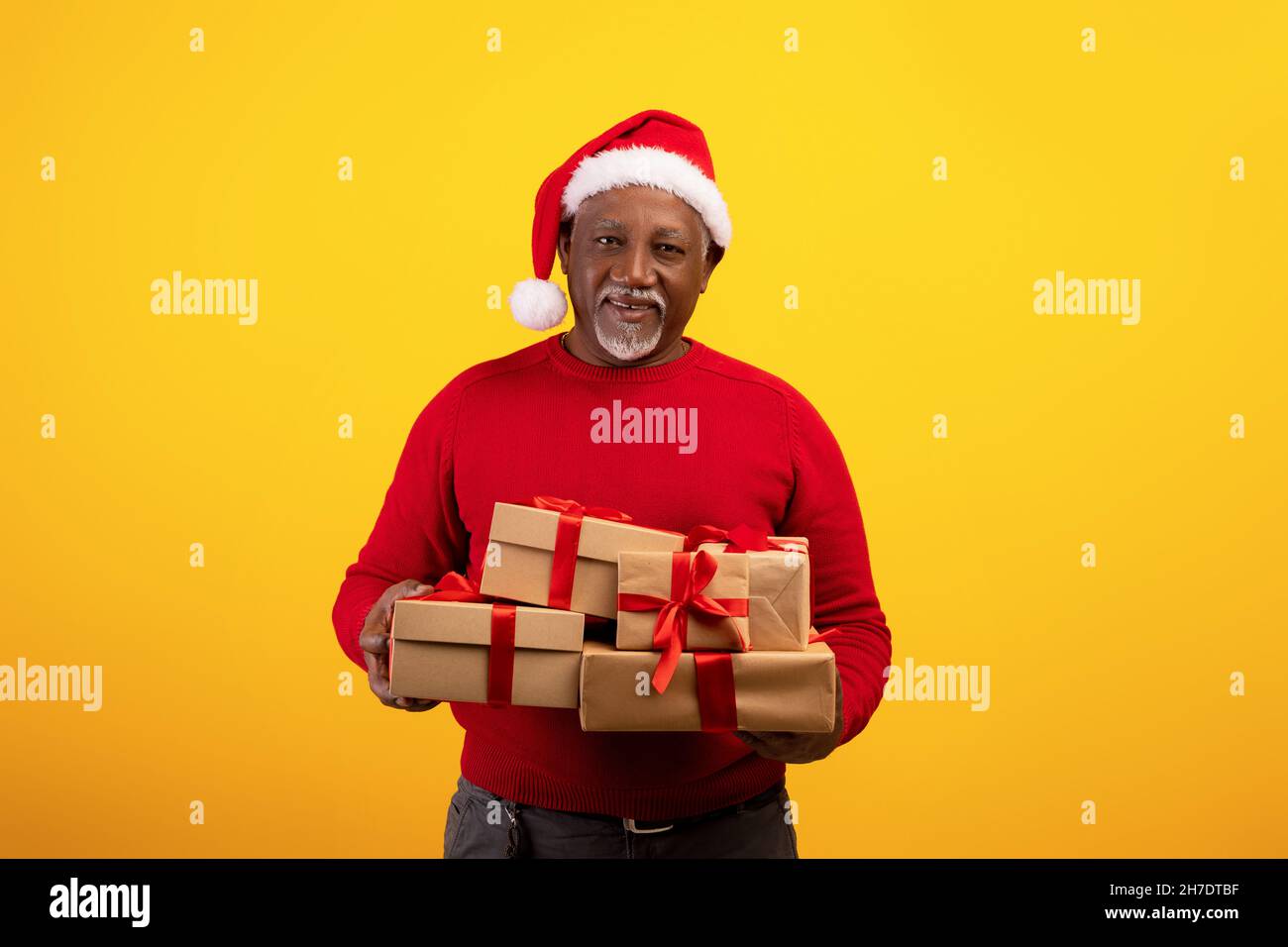 Positive senior black man holding heap of wrapped Xmas gift boxes, wearing Santa hat on orange studio background Stock Photo