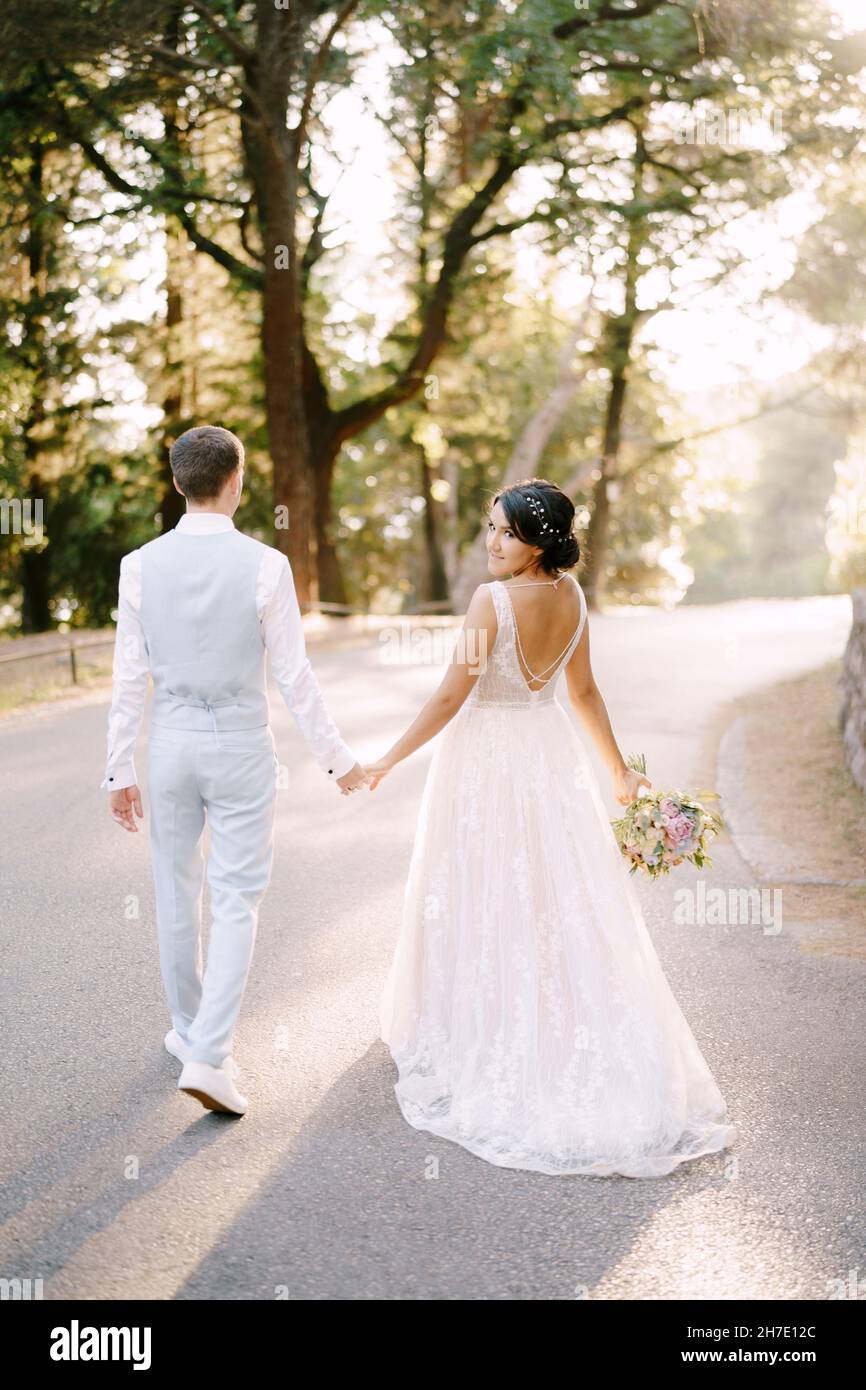 The bride and walk holding hands on the road among the trees in an ...