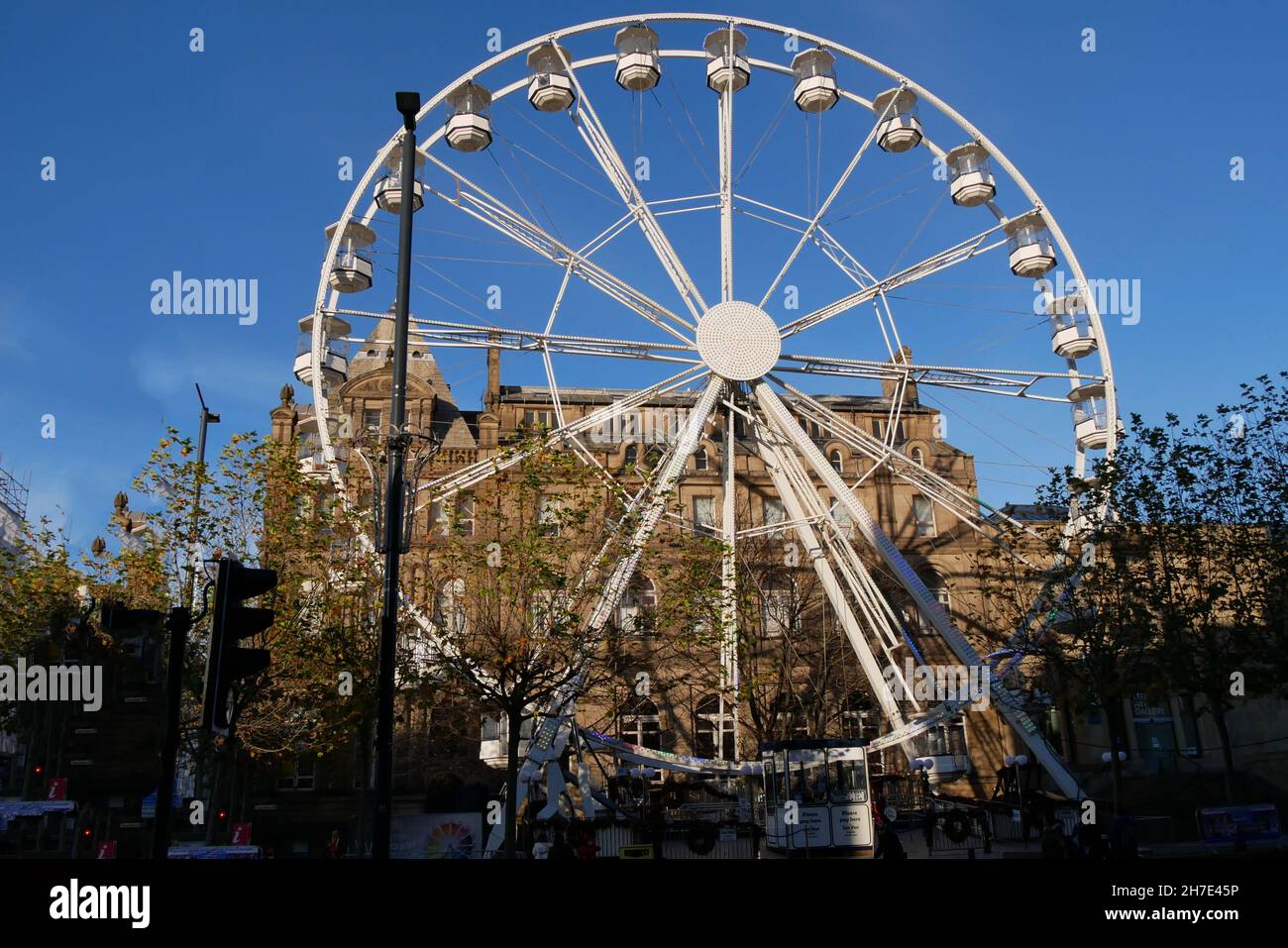 Large white ferris wheel with multiple viewing pods in front of large stone building small trees in foreground and blue sky Stock Photo