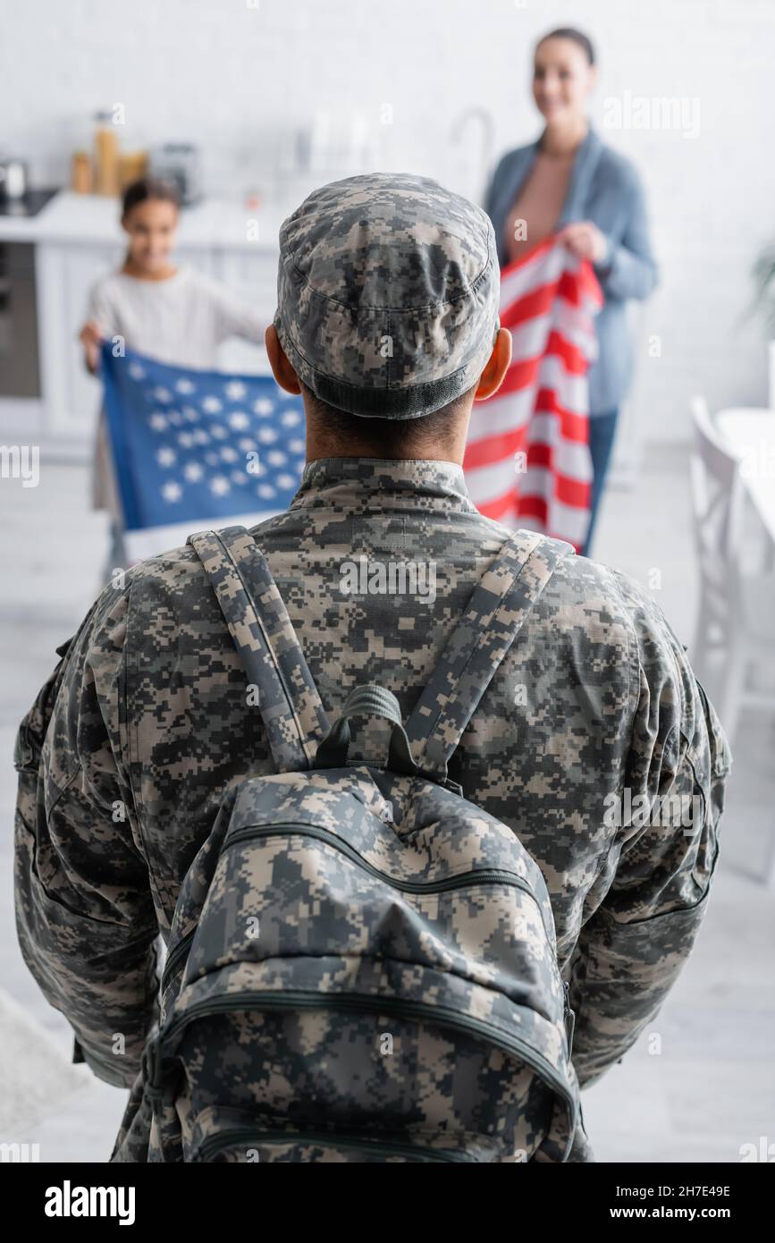 Man in camouflage uniform with backpack near blurred family with american flag at home Stock Photo