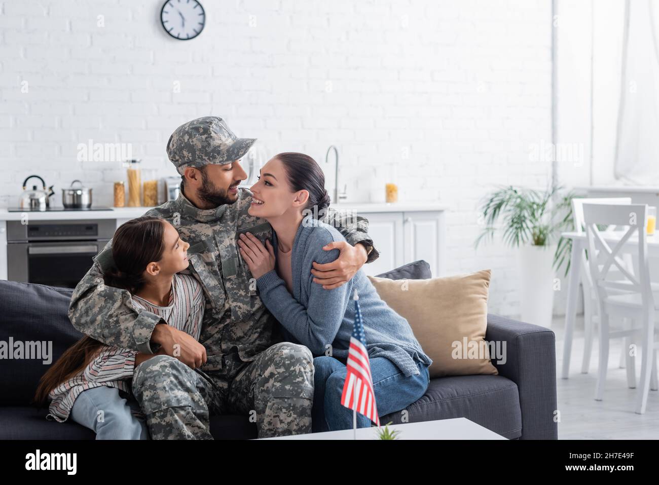 Man in camouflage uniform hugging family near american flag at home Stock Photo