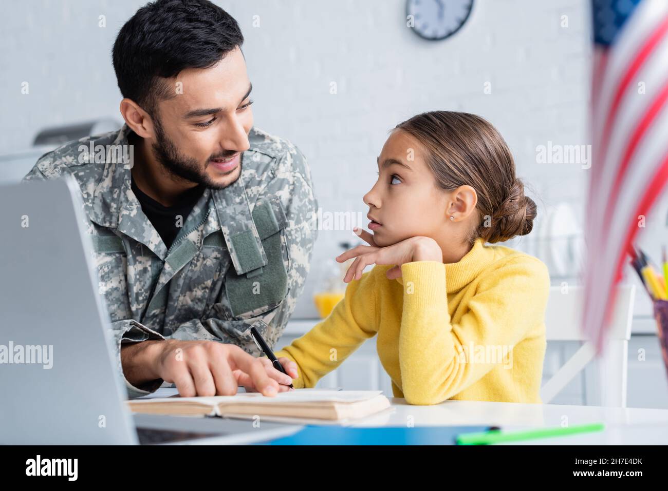 Smiling father in camouflage uniform pointing at book near daughter, laptop and american flag at home Stock Photo