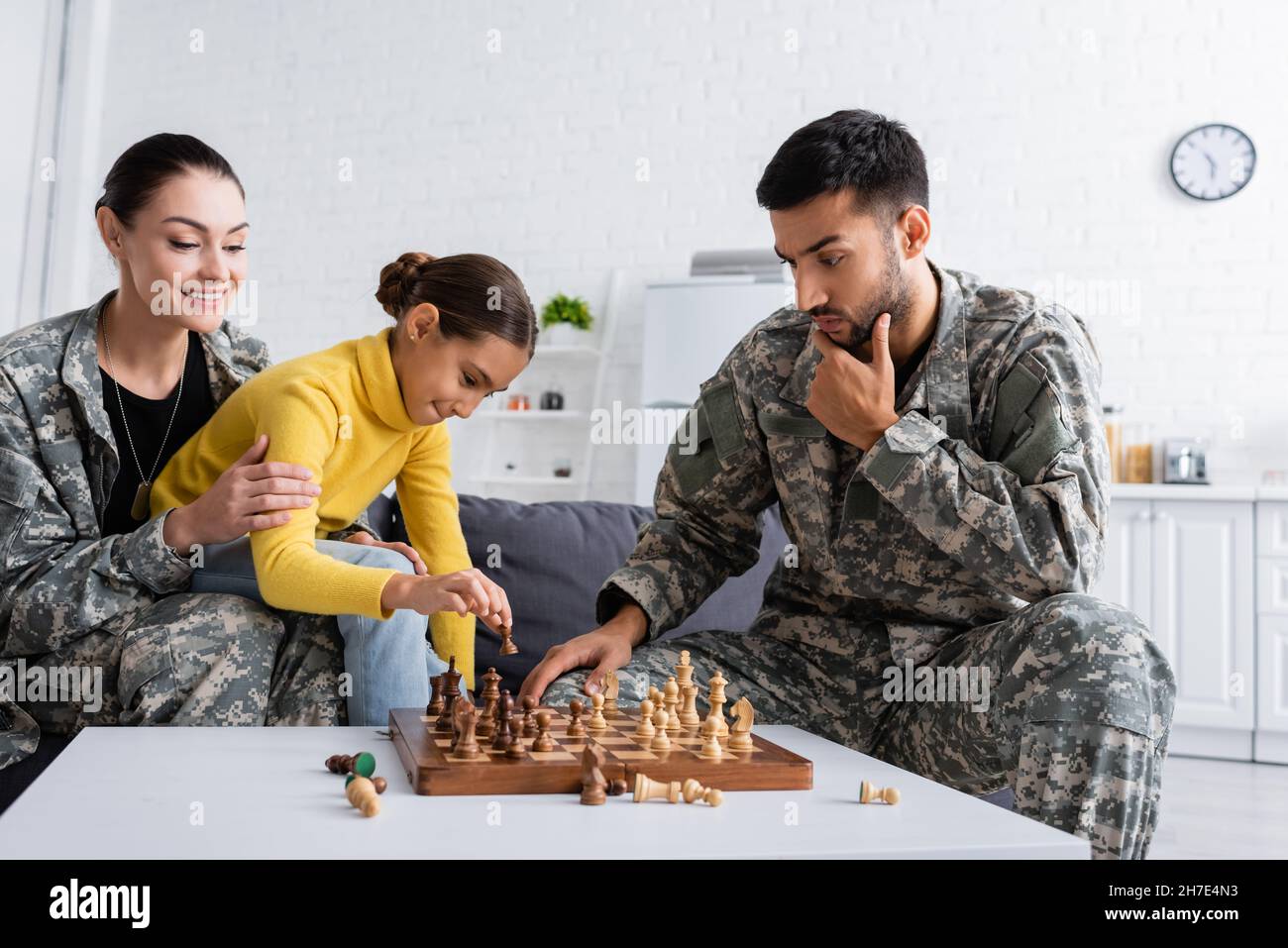 Pensive father in camouflage uniform playing chess near smiling mom and kid at home Stock Photo