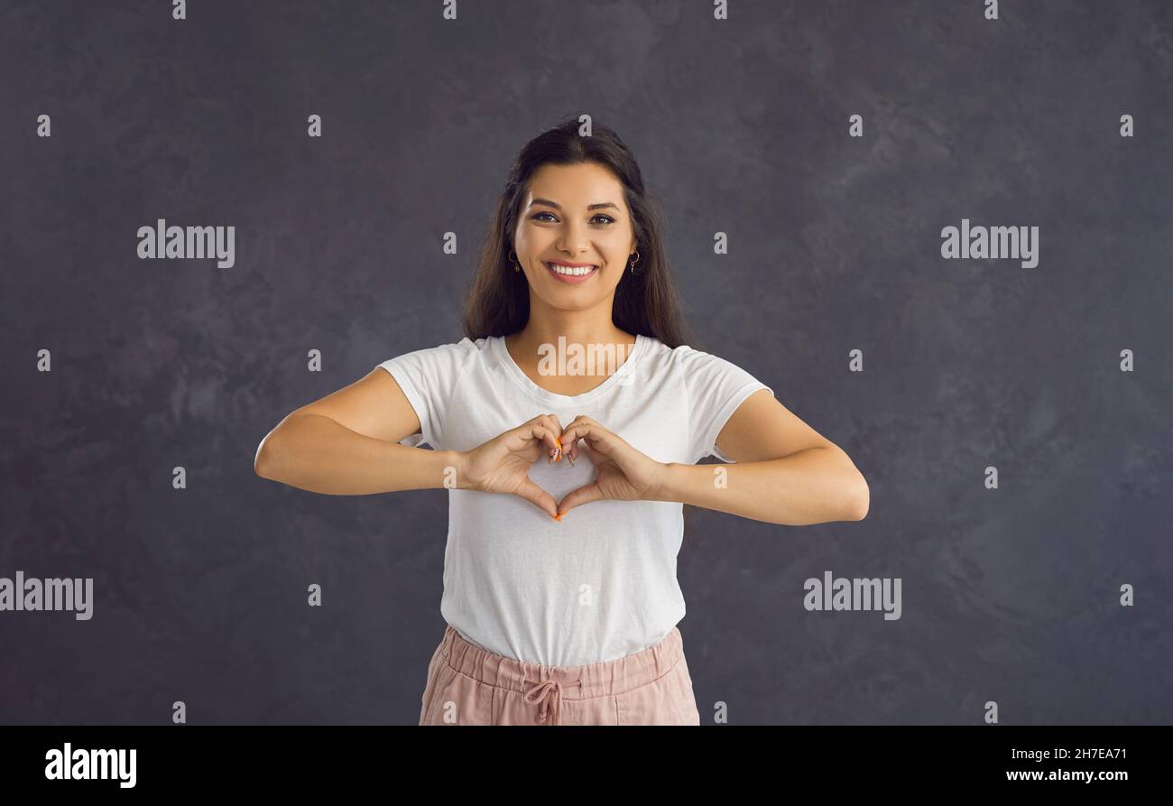 Portrait of a young smiling woman showing a gesture of love standing on a gray background. Stock Photo