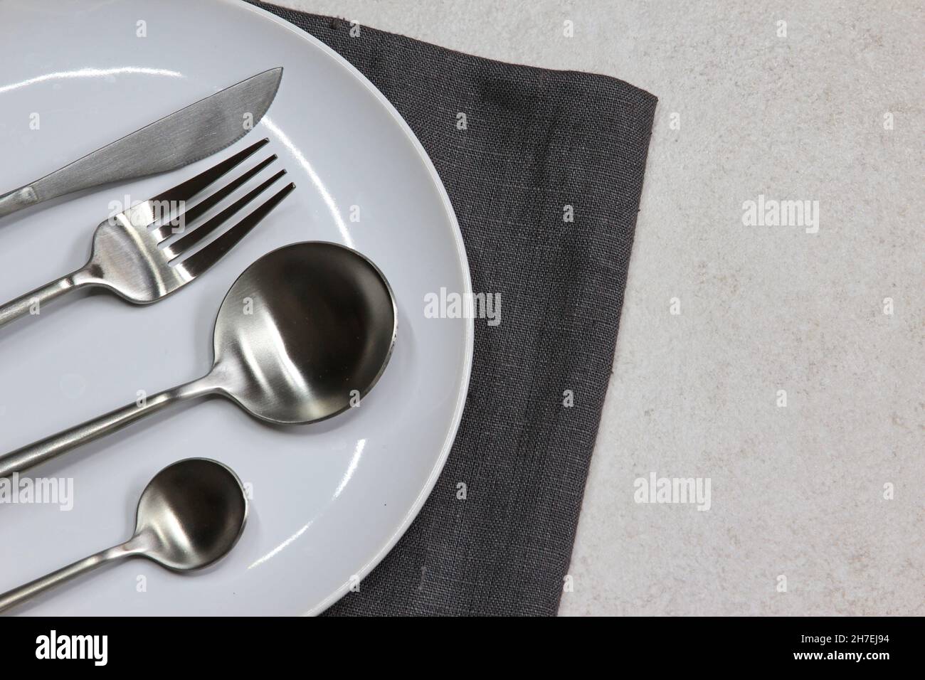 White empty plate with knife, spoons and fork dark textile napkin on ...