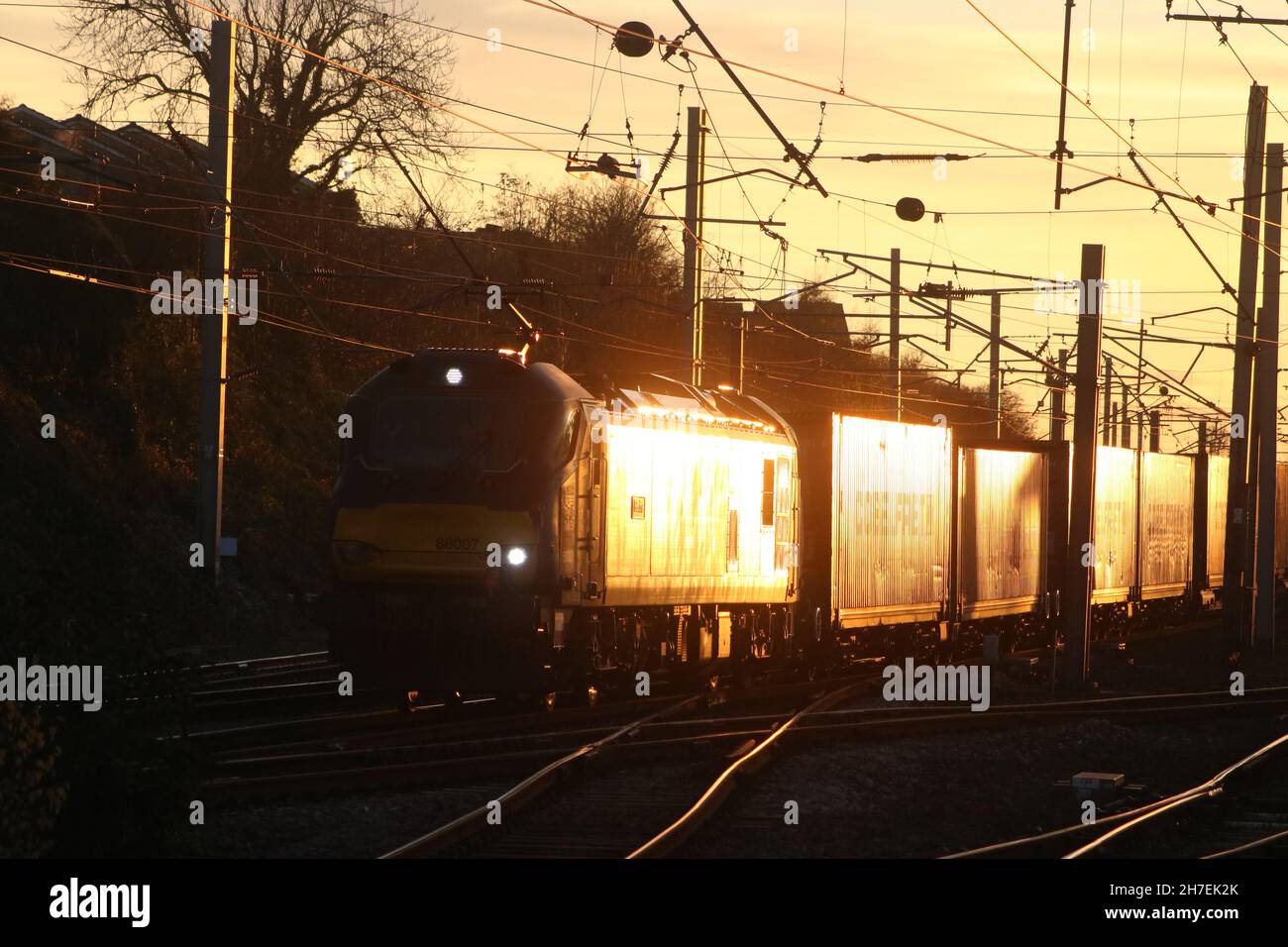 Sunset glint reflecting off electric loco and containers as freight train passes through Carnforth on West Coast Main line on 22nd November 2021. Stock Photo