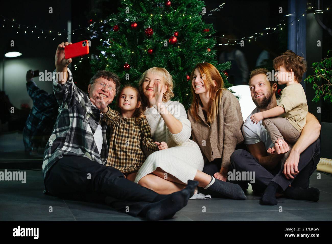Sitting on the floor big family posing for a christmas selfie. Children, parents and grandparents sitting in front of a decorated christmas tree. Stock Photo