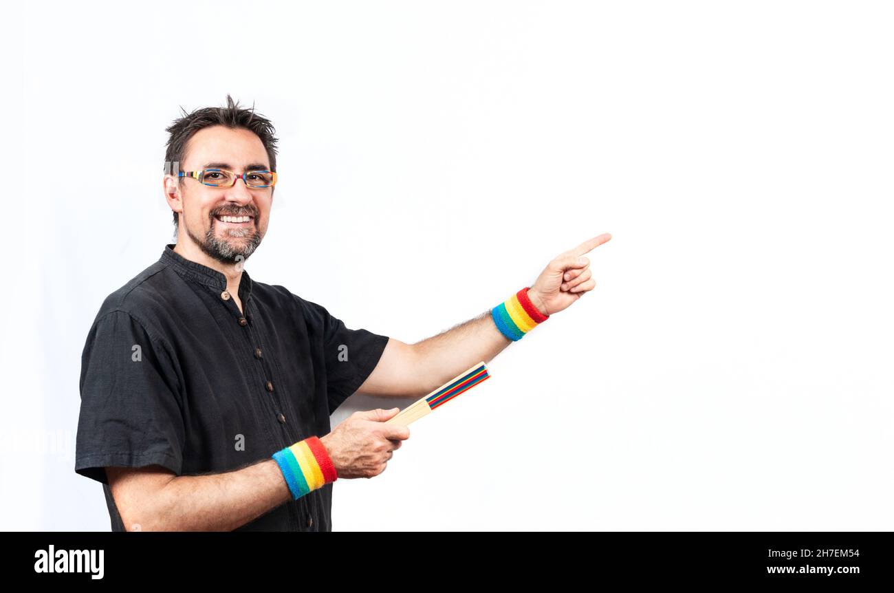 Man in black shirt wearing rainbow colored glasses and wristbands; smiling at the camera and pointing to a blank space with one hand and a closed fan Stock Photo