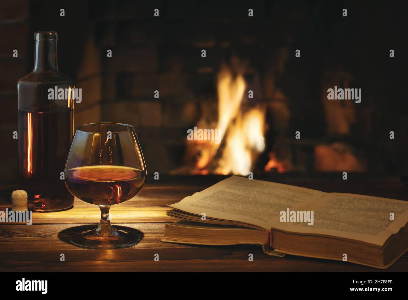 Glass of cognac, a bottle and an open old book on the table near the burning fireplace. Rest and relaxation concept Stock Photo