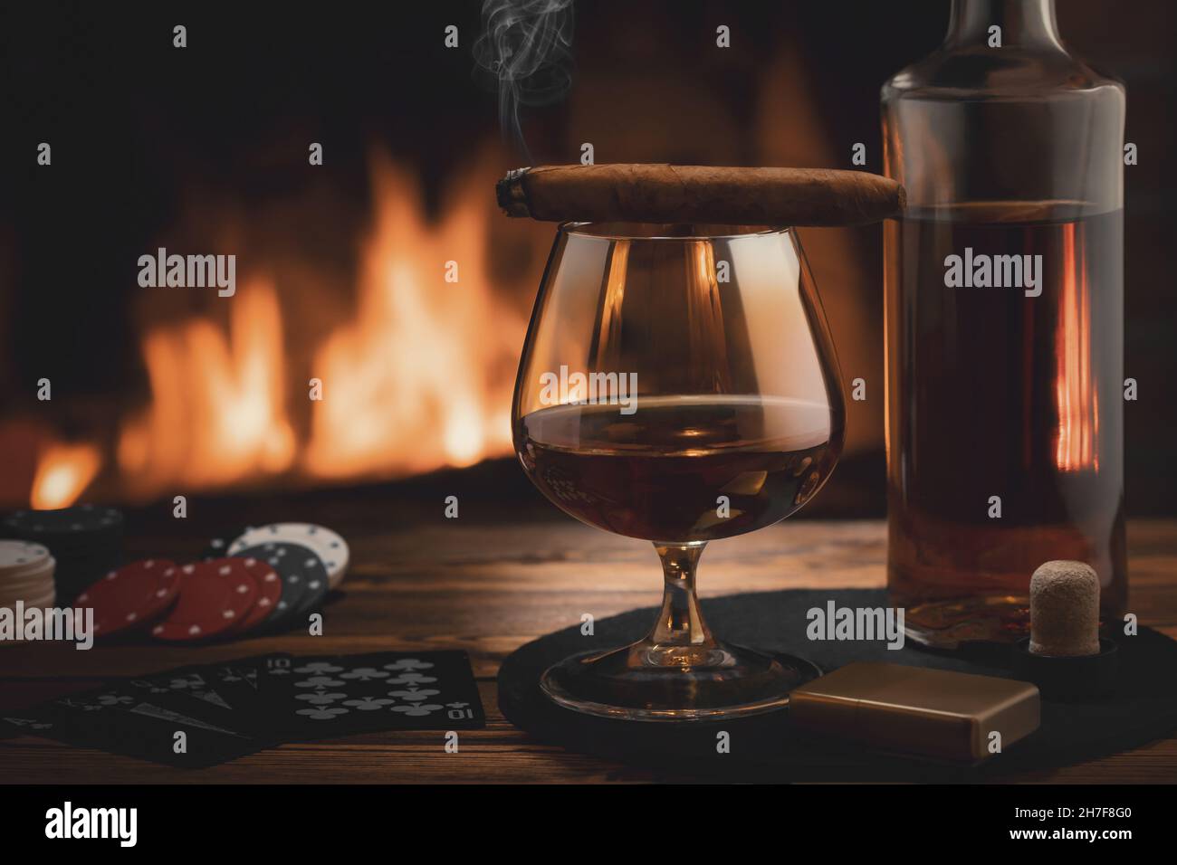 Glass of cognac, cigar, bottle, cards game and chips on the table near the burning fireplace. Rest, pleasure, good luck and dolce vita concept. Stock Photo