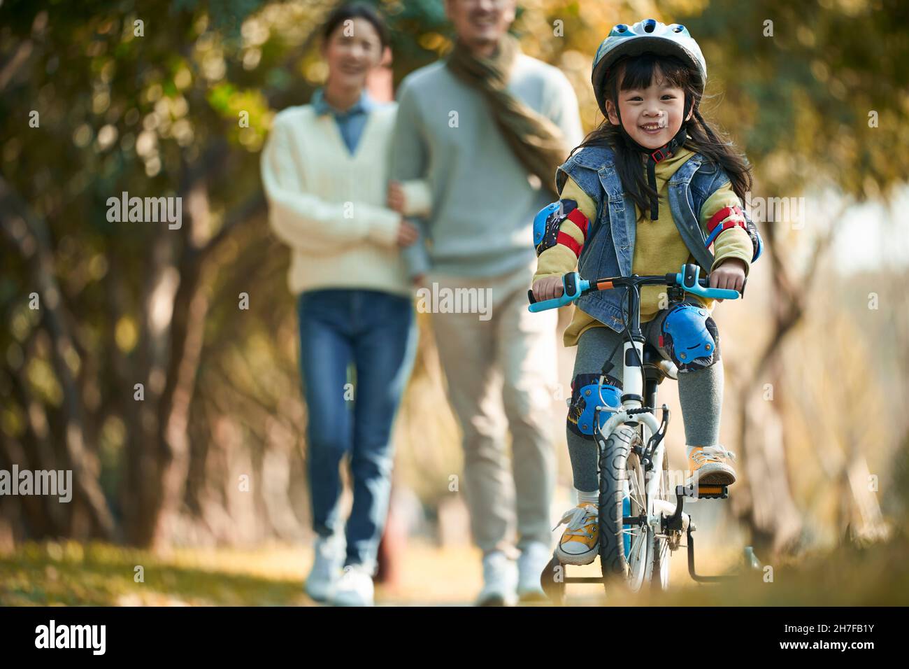 little asian girl with helmet and full protection gears riding bike in city park with parents watching from behind Stock Photo