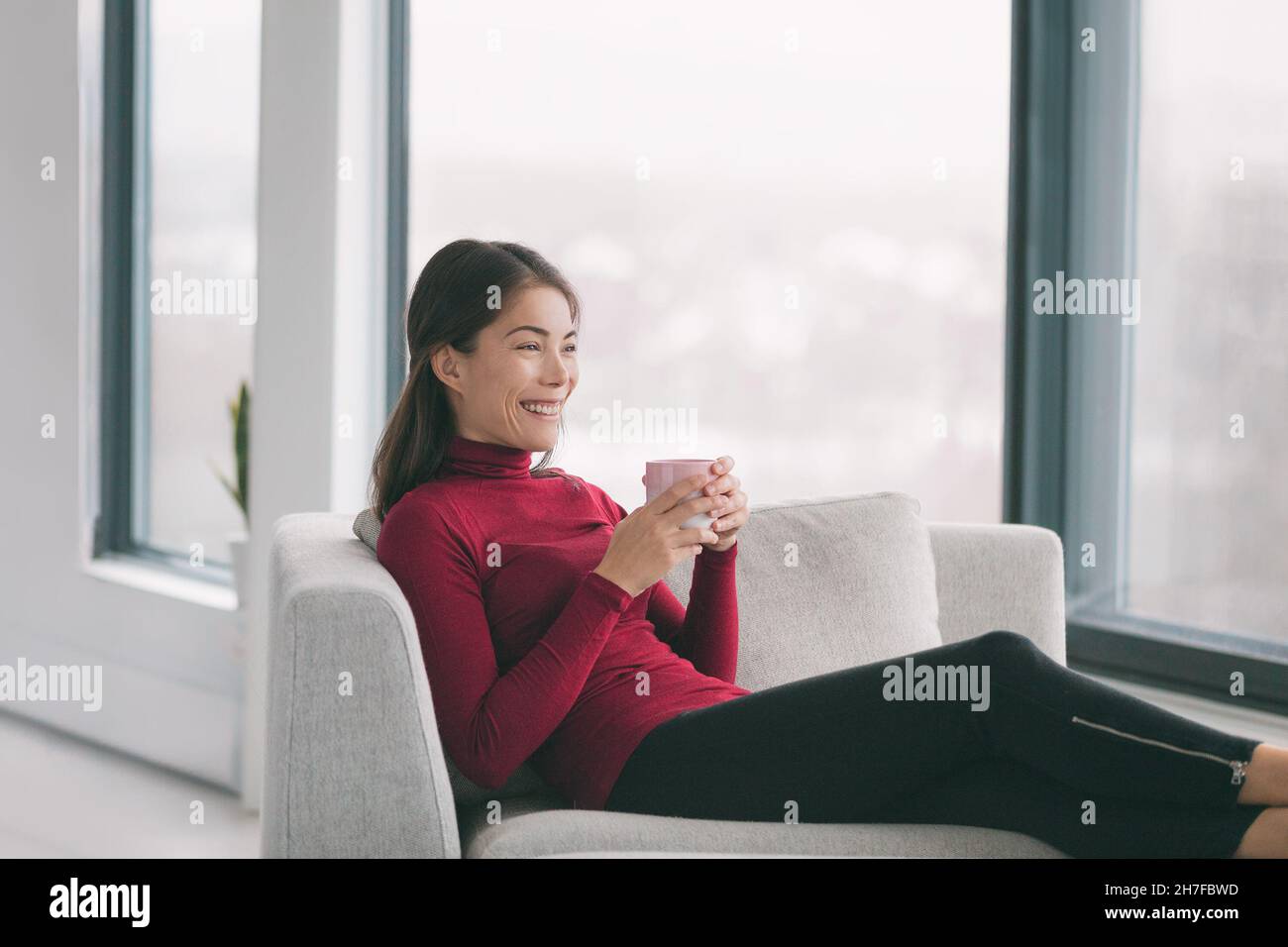Home lifestyle Happy Asian woman relaxing drinking tea in living room enjoying free time on weekend to relax at window view sitting in sofa. Indoor Stock Photo
