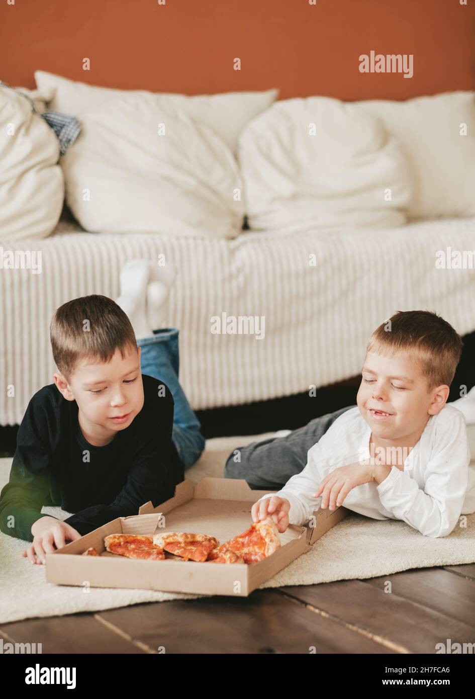 two boys lie on the floor eating pepperoni pizza from a box. vertical frame. two boys lie on the floor eating pepperoni pizza from a box. Stock Photo