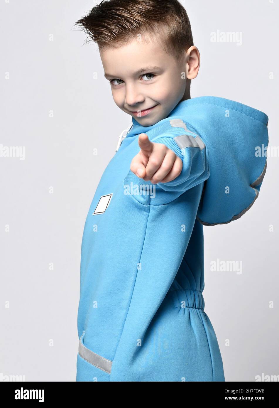 Portrait of tricky smiling kid boy in blue jumpsuit with hood standing sideways looking and pointing finger at camera Stock Photo