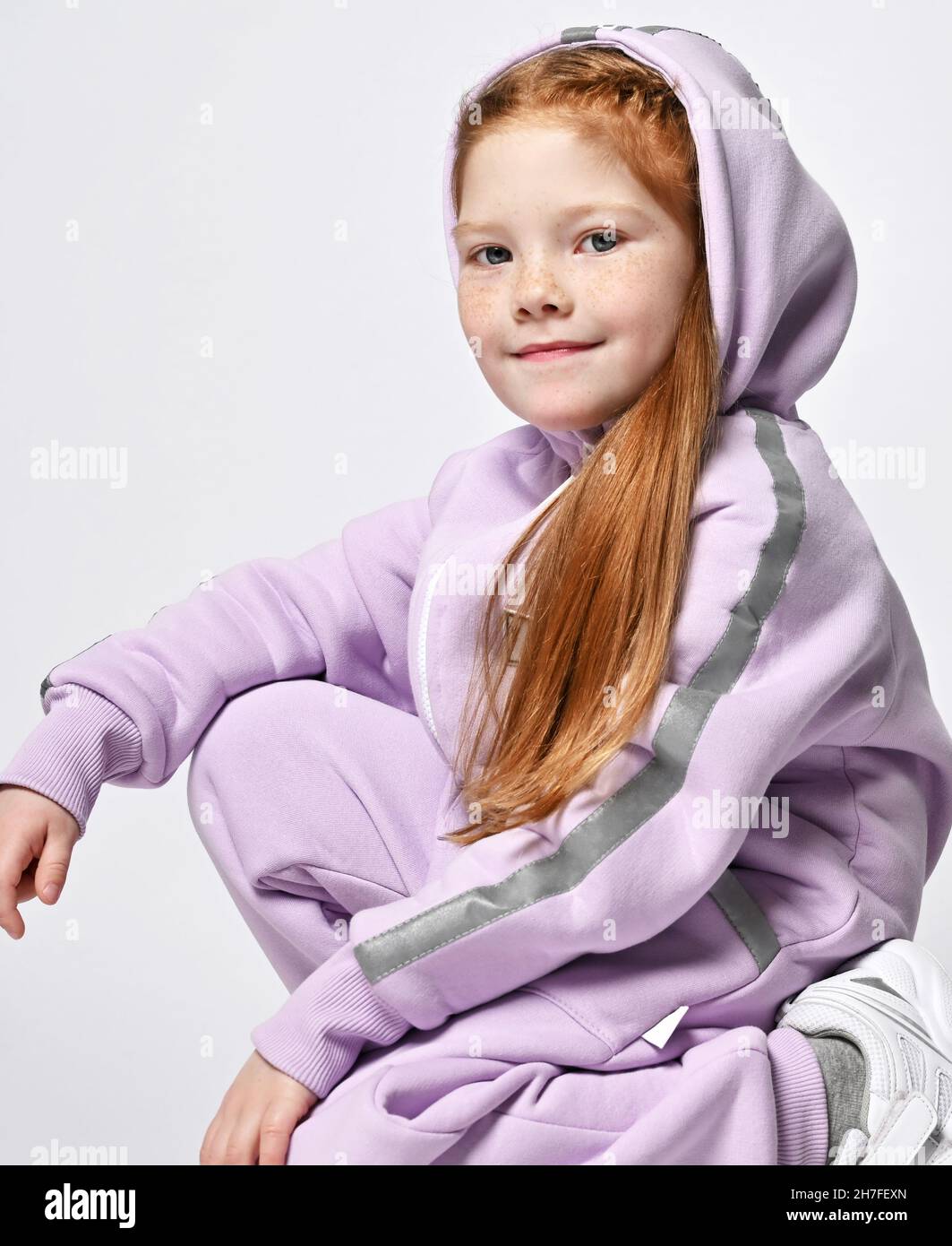 Portrait of cool red-haired kid girl in pink jumpsuit sitting squatted sideways to camera with her hood on her head Stock Photo