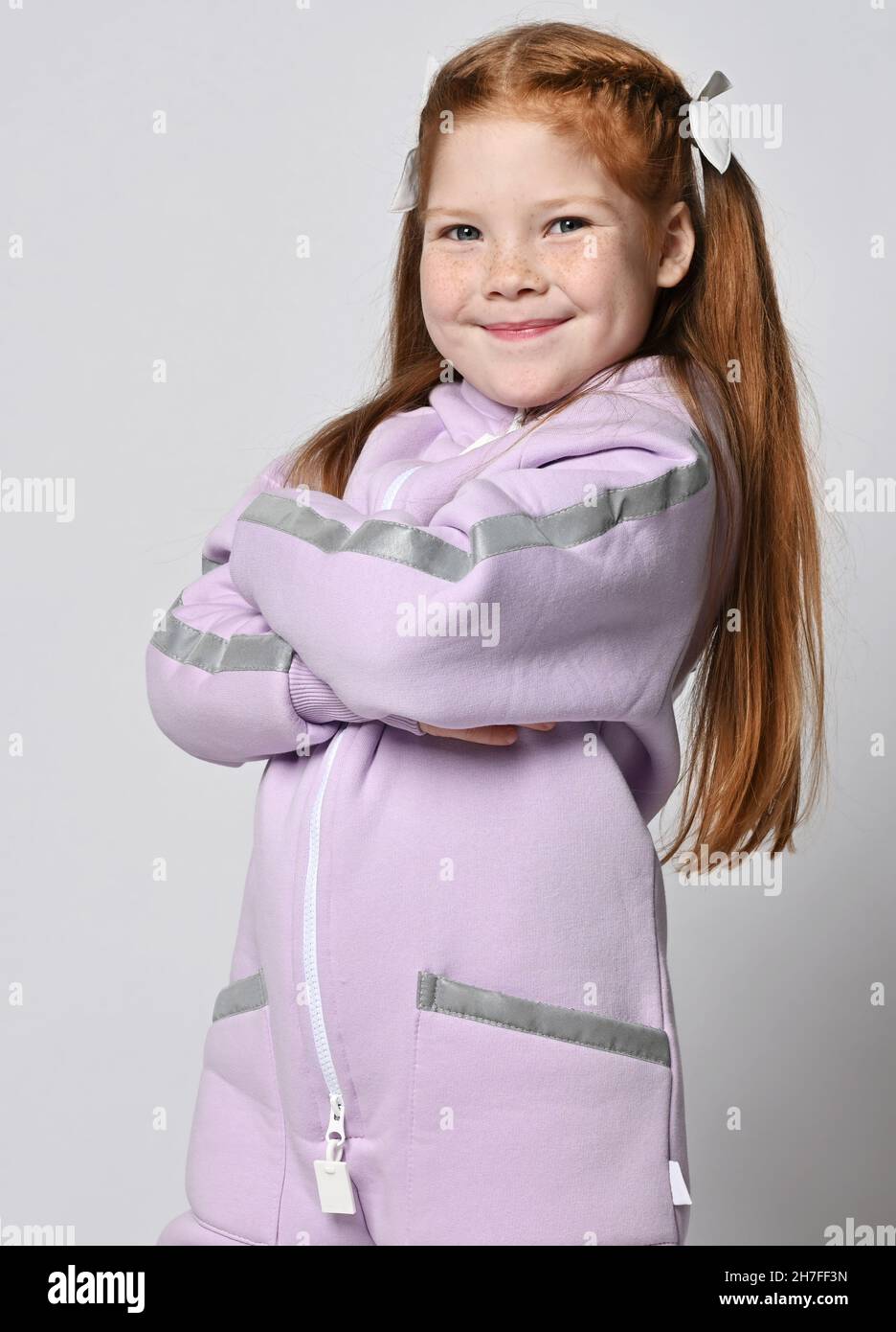 Portrait of cute smiling kid girl in pink modern jumpsuit standing with arms crossed at chest, looking at camera Stock Photo