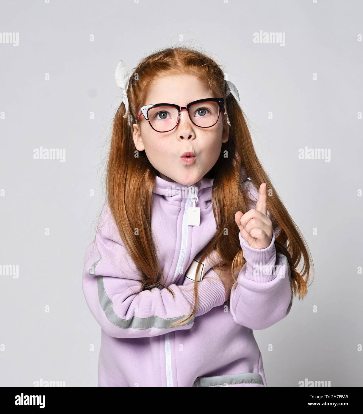 Portrait of smart redhead kid girl in glasses and pink jumpsuit with hood standing holding finger up, got an idea Stock Photo