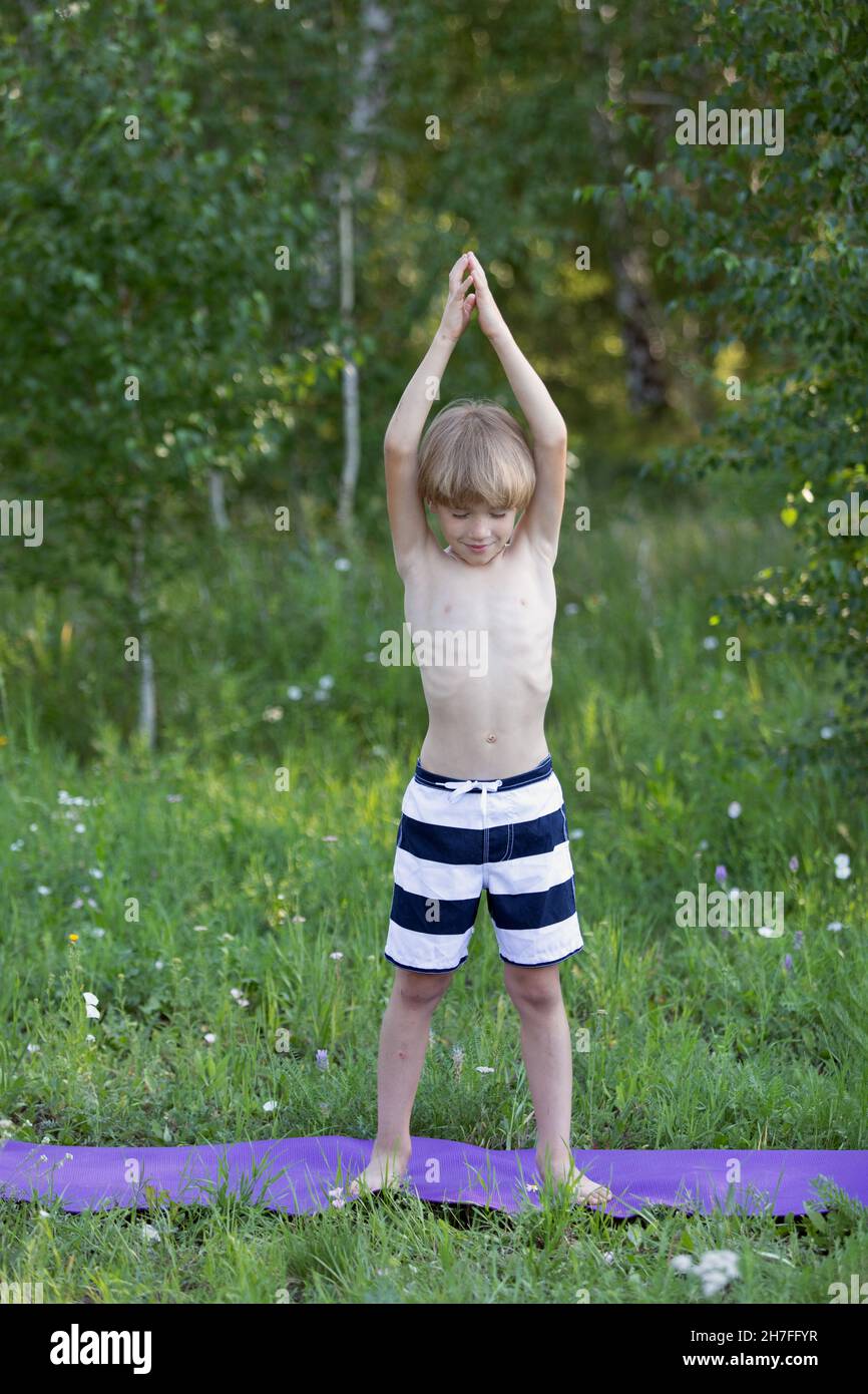 Boy practising yoga outdoor in green park, performs variant of mountain pose, with his hands raised, called asana Talasana Stock Photo