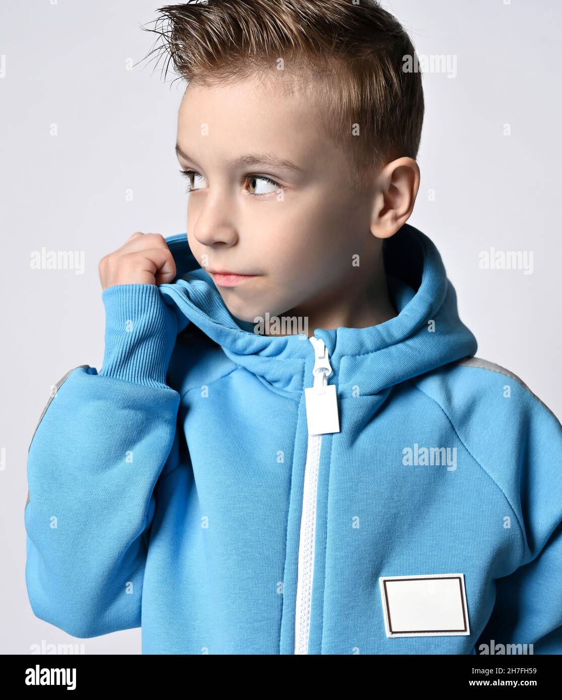 Portrait of kid boy in blue jumpsuit or hoodie with zipper standing with head turned, looking aside holding his cowl Stock Photo
