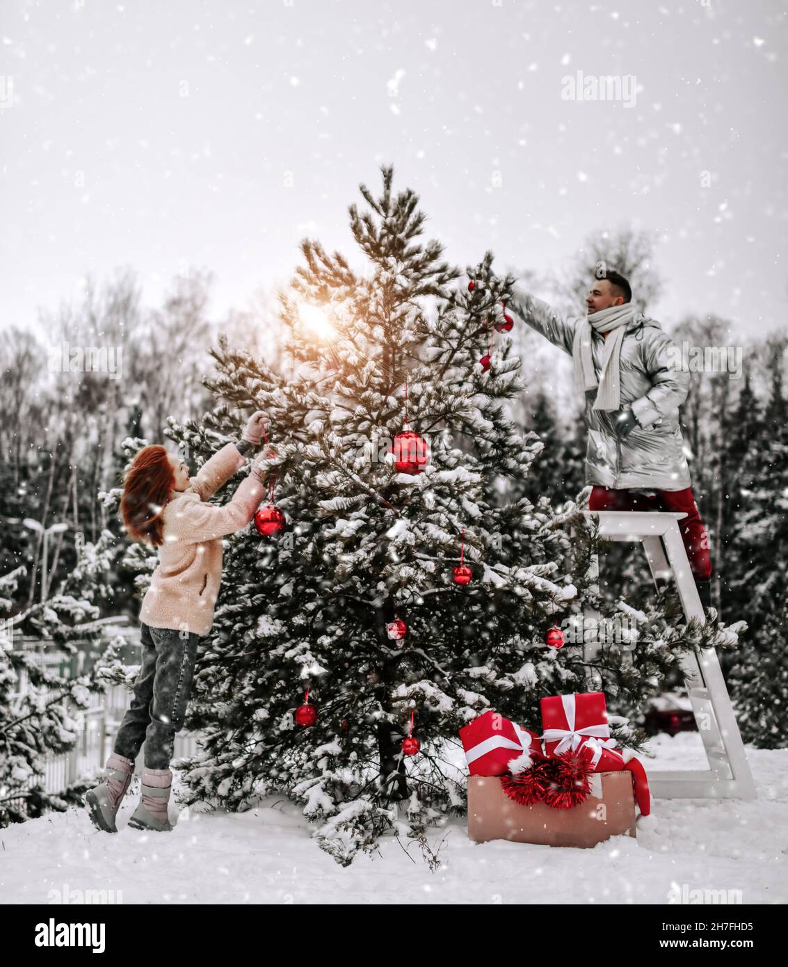 Young happy couple in stylish winter clothing decorating Christmas tree with balls outdoors in winter snowy forest Stock Photo