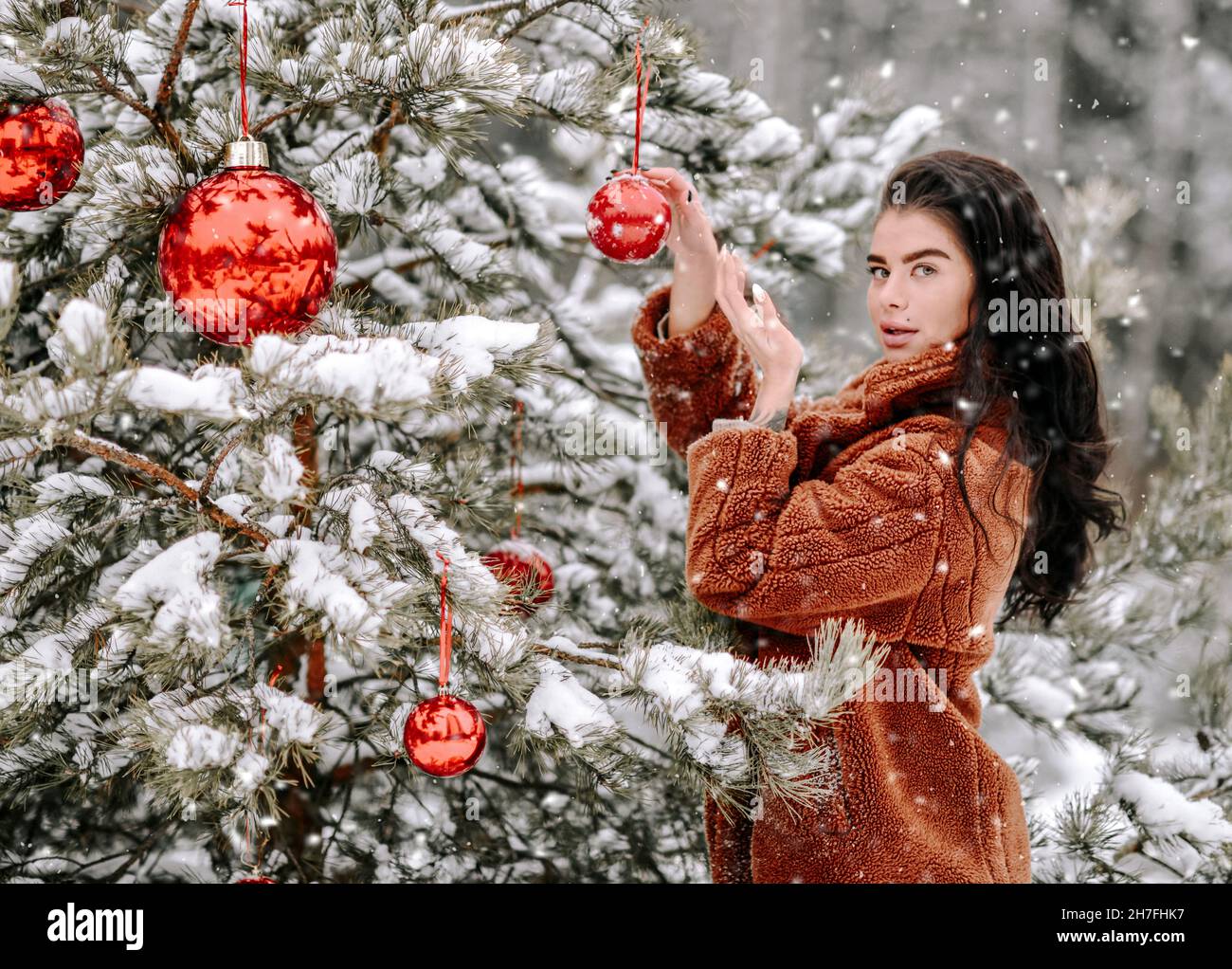 Young sexy woman in stylish fur coat stands near decorates Christmas tree with red balls under snow Stock Photo