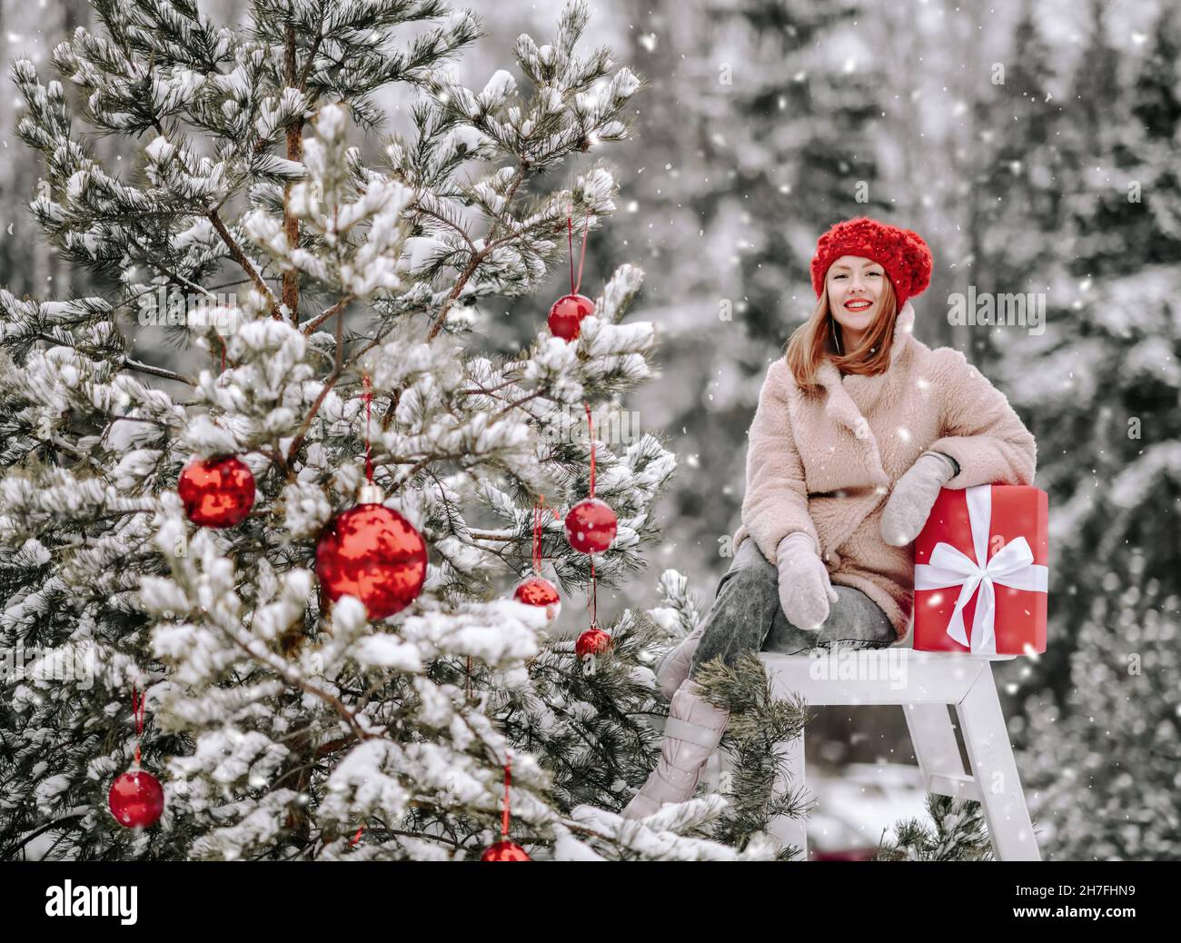 Young smiling pretty woman in stylish warm clothing sitting with holiday gift on stool near decorated Christmas outdoors Stock Photo