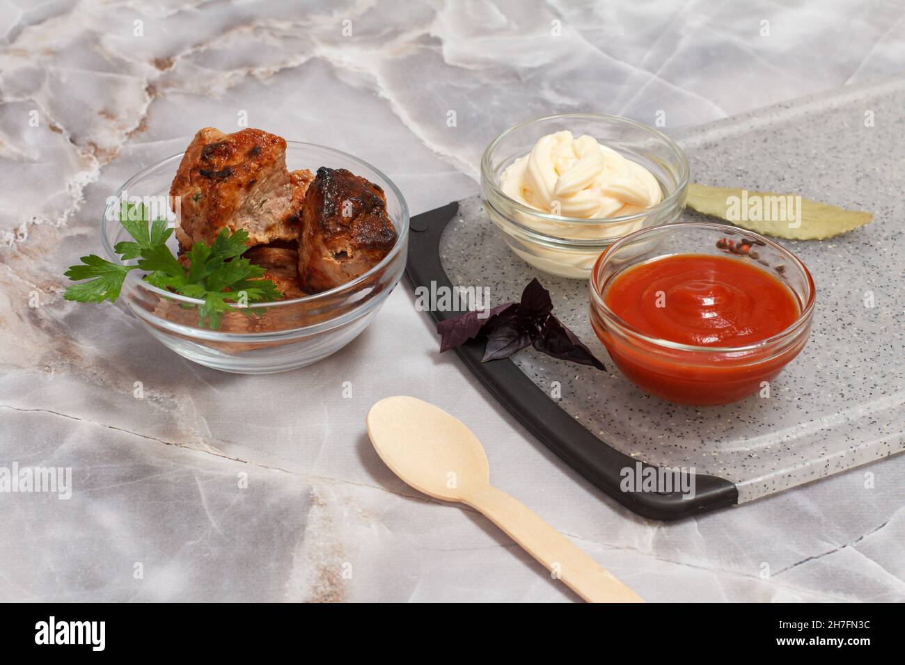 Grilled pork in a glass bowl with fresh basil. Porcelain bowls with ketchup and cheese sauce on the cutting board. Top view. Stock Photo