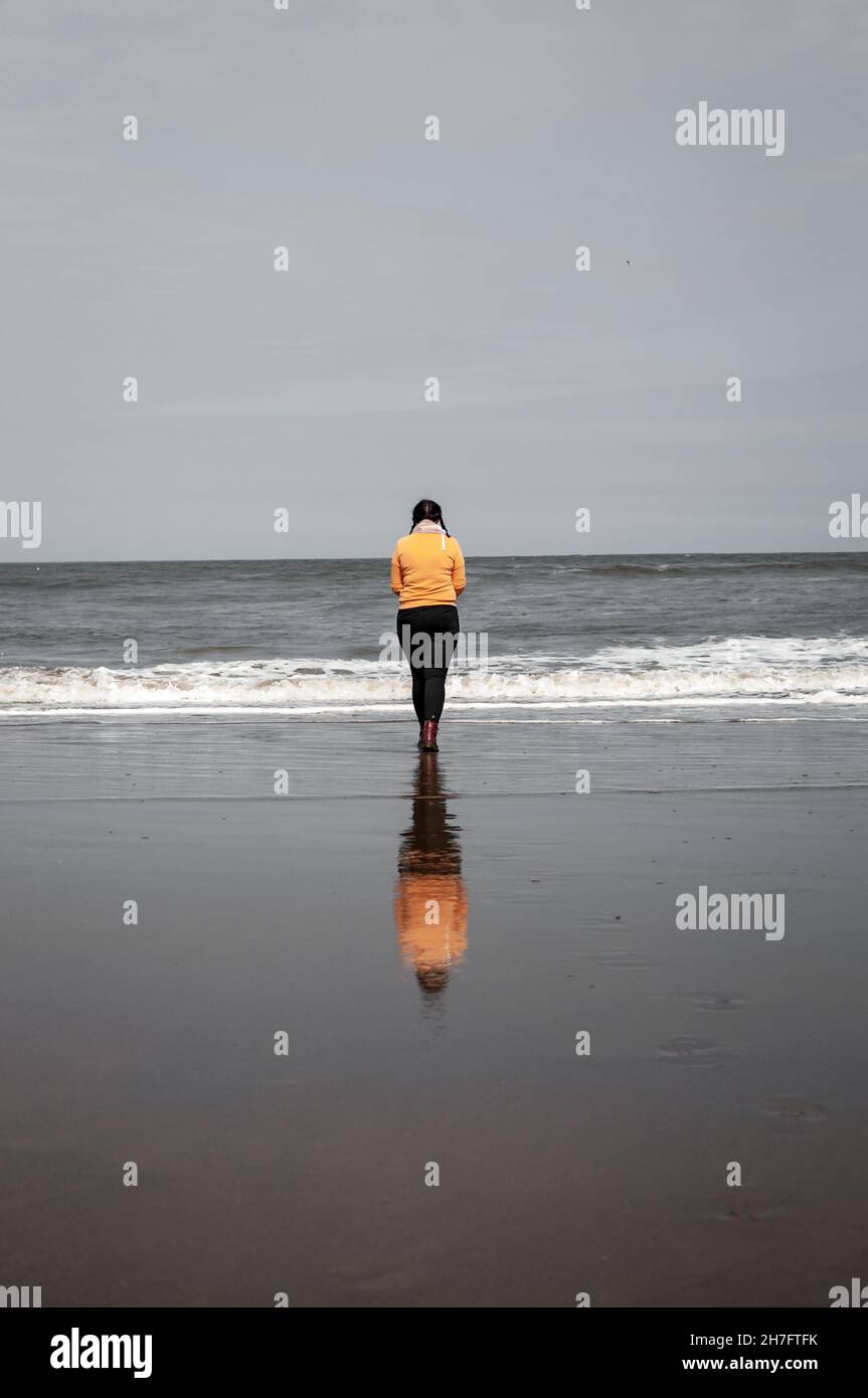 A lone figure stands at the edge of the ocean, her reflection cast upon the sand Stock Photo