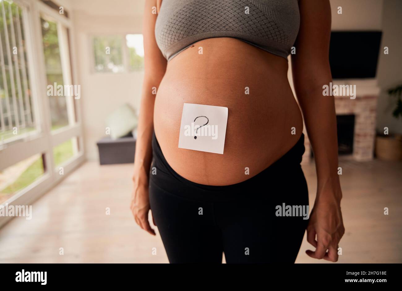Caucasian pregnant woman standing in living room with question mark on belly in living room Stock Photo