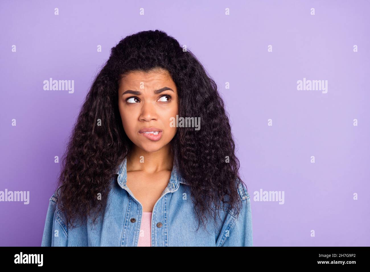 Photo of doubtful cute dark skin woman dressed jeans shirt biting lip looking empty space isolated violet color background Stock Photo