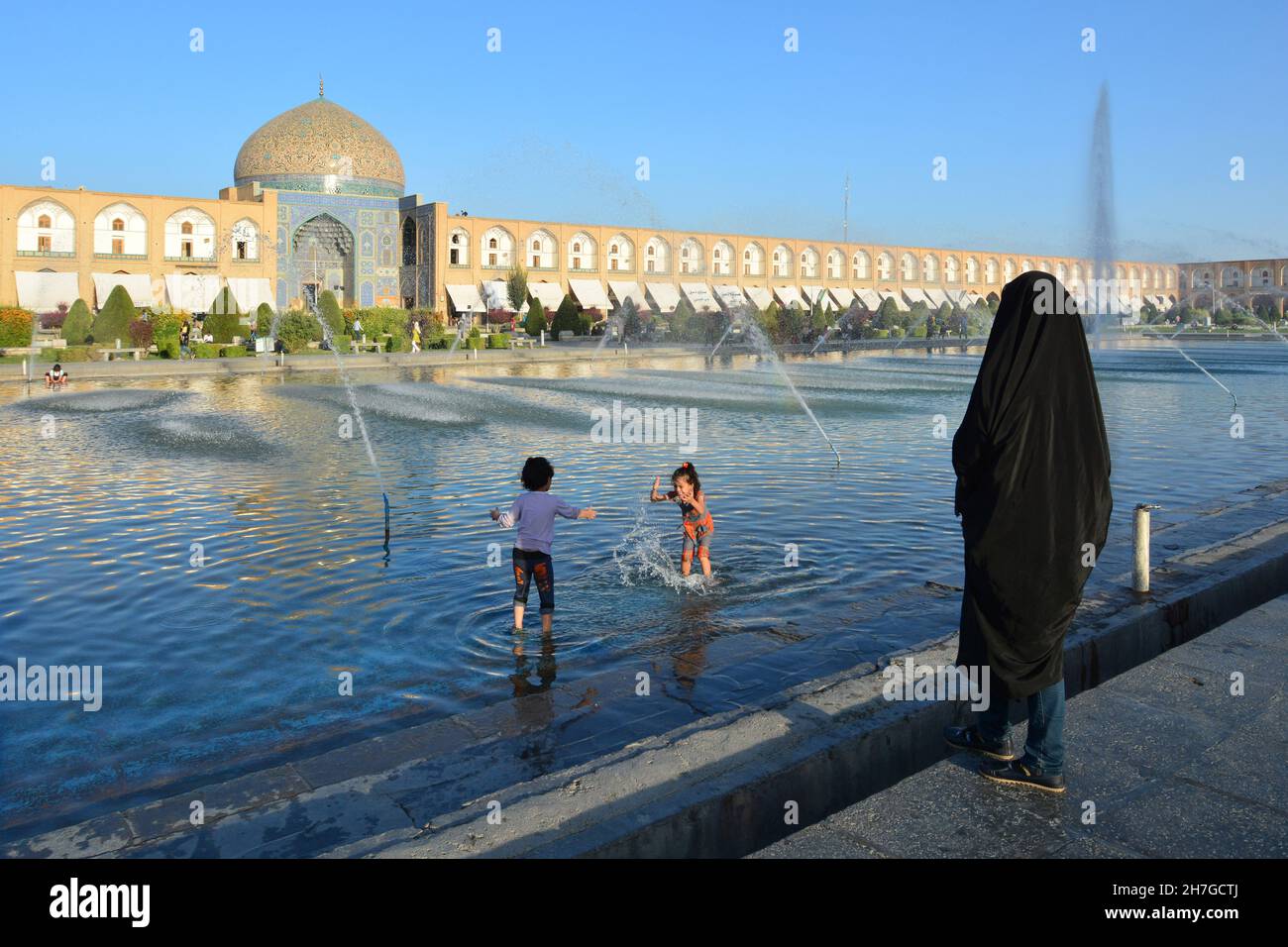 IRAN. ISPAHAN ESFAHAN. EMAM SQUARE. WOMAN IN VEIL LOOKING HER KIDS PLAYING WITH THE WTAER IN THE FOUNTAIN FACING THE CHEICK LOFTOLLAH MOSQUE. Stock Photo