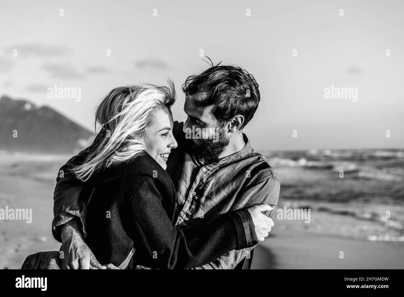 Romantic happy couple in love hugging each other on shoreline at the wild beach - Boyfriend and girlfriend smiling in front of the sea during a big hu Stock Photo