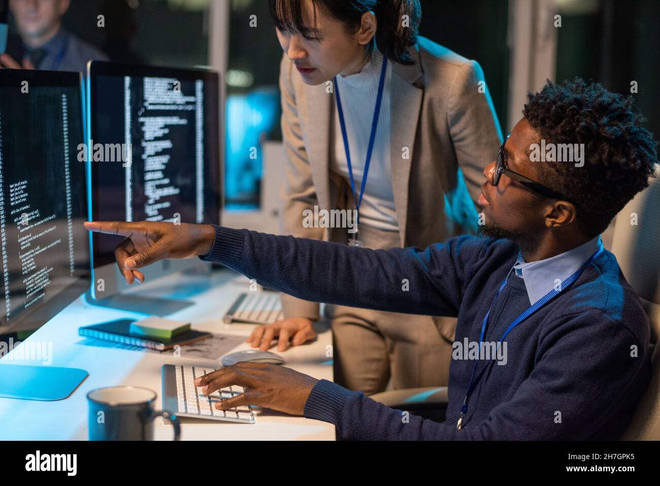 Two young intercultural colleagues discussing coded data on computer screen while African businessman pointing at it Stock Photo