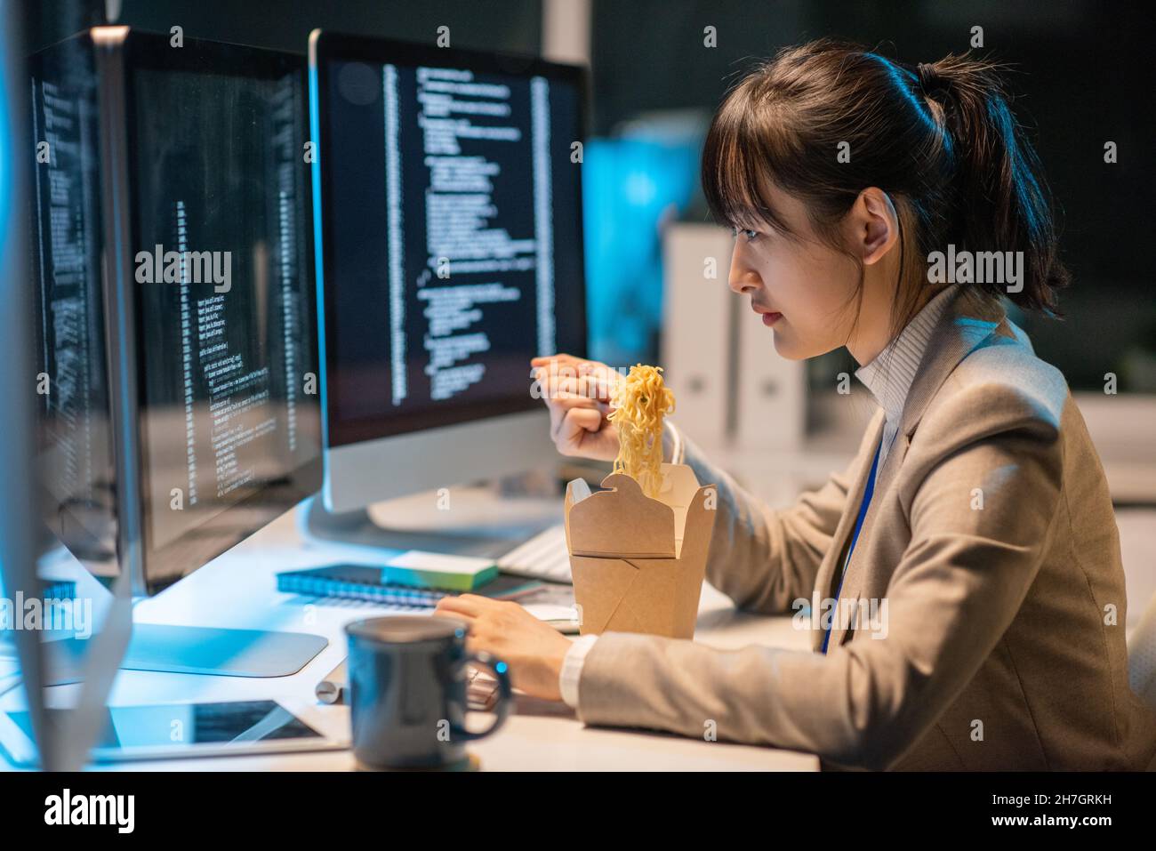 Side view of young Asian female programmer eating chinese wok during work over coded data in front of computer monitor Stock Photo