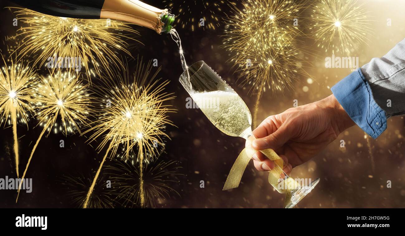 Filling champagne glass adorned with gold bow from a bottle with fireworks background at a party. Front view. Stock Photo