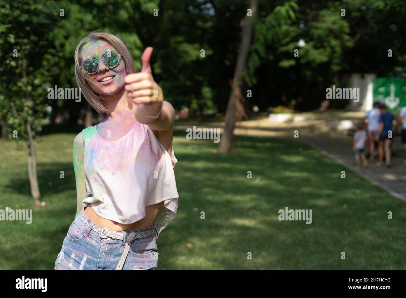 happy young woman at Holi festival. Traditional Indian festival. Blonde in the park with a face in colorful powder paint. Shows the class with fingers Stock Photo