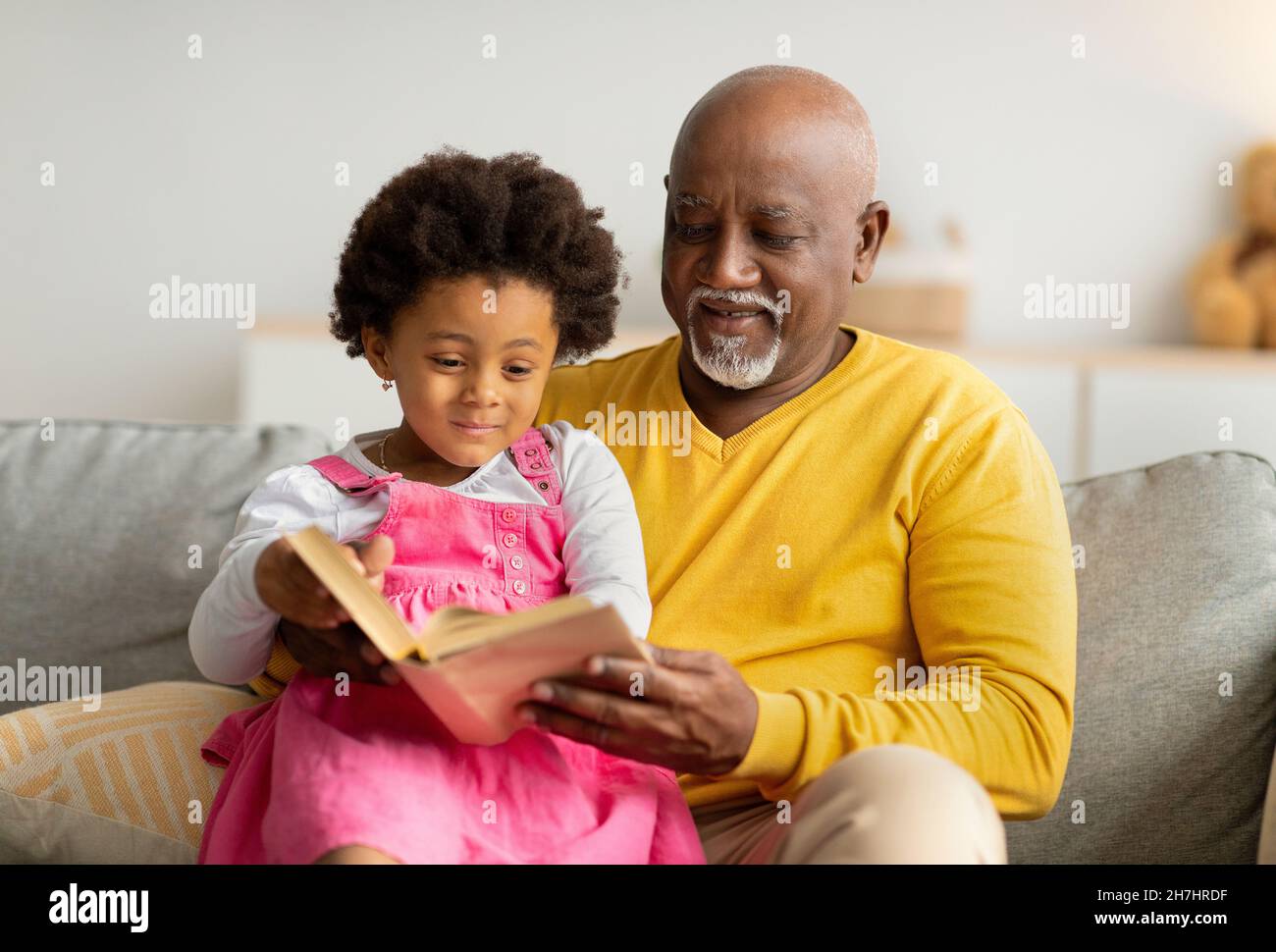 Satisfied black little kid and elderly man reading book with fairy tales on sofa in living room interior Stock Photo
