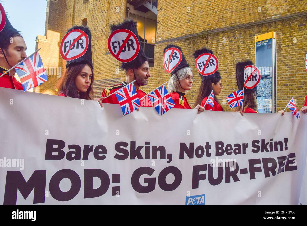 London, UK. 23rd Nov, 2021. Activists wearing guard costumes and caps with anti-fur signs hold Union Jack flags and a banner asking the Ministry of Defence (MOD) to go fur-free, during the demonstration.PETA activists partially stripped off their clothing next to Tower Bridge, calling on the UK government and the Ministry of Defence to use faux fur in guards' caps instead of real bearskin. The demonstration also marked the 21st anniversary of the fur-farming ban in England and Wales. Credit: SOPA Images Limited/Alamy Live News Stock Photo